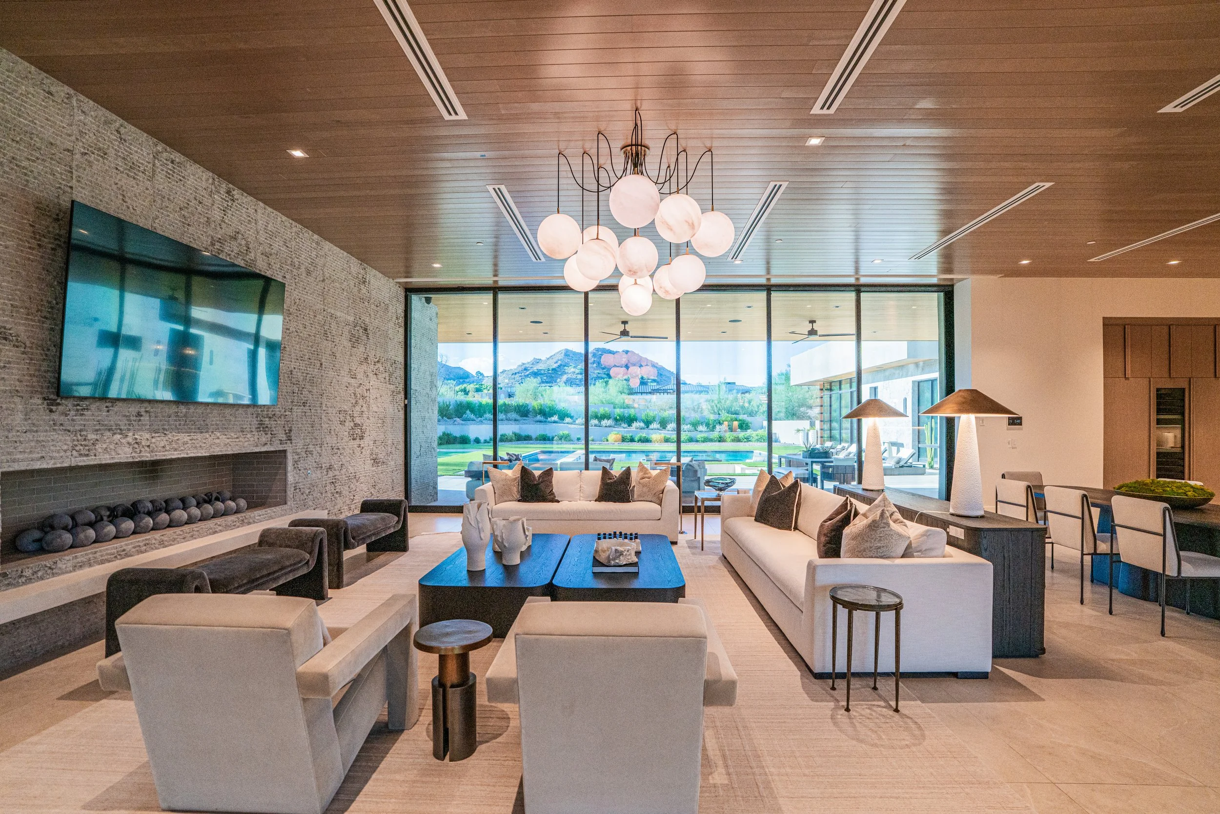 A spacious modern living room with white sofas, dark coffee table, and armchairs. Large floor-to-ceiling windows offer a view of mountains and greenery outside. A chandelier with round white globes hangs from a wooden ceiling. The room features a bri