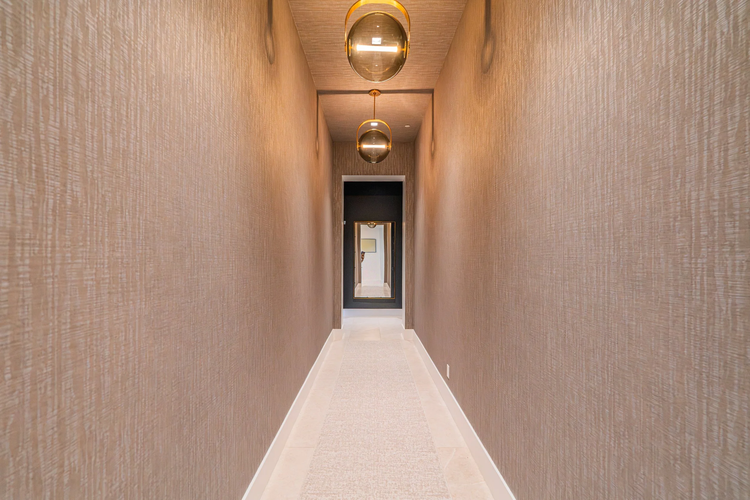 A long, narrow hallway with beige textured walls beautifully lit by three hanging spherical gold and glass pendant lights. At the end, there is a black-framed mirror reflecting part of the hallway and an open door leading to another room with a white