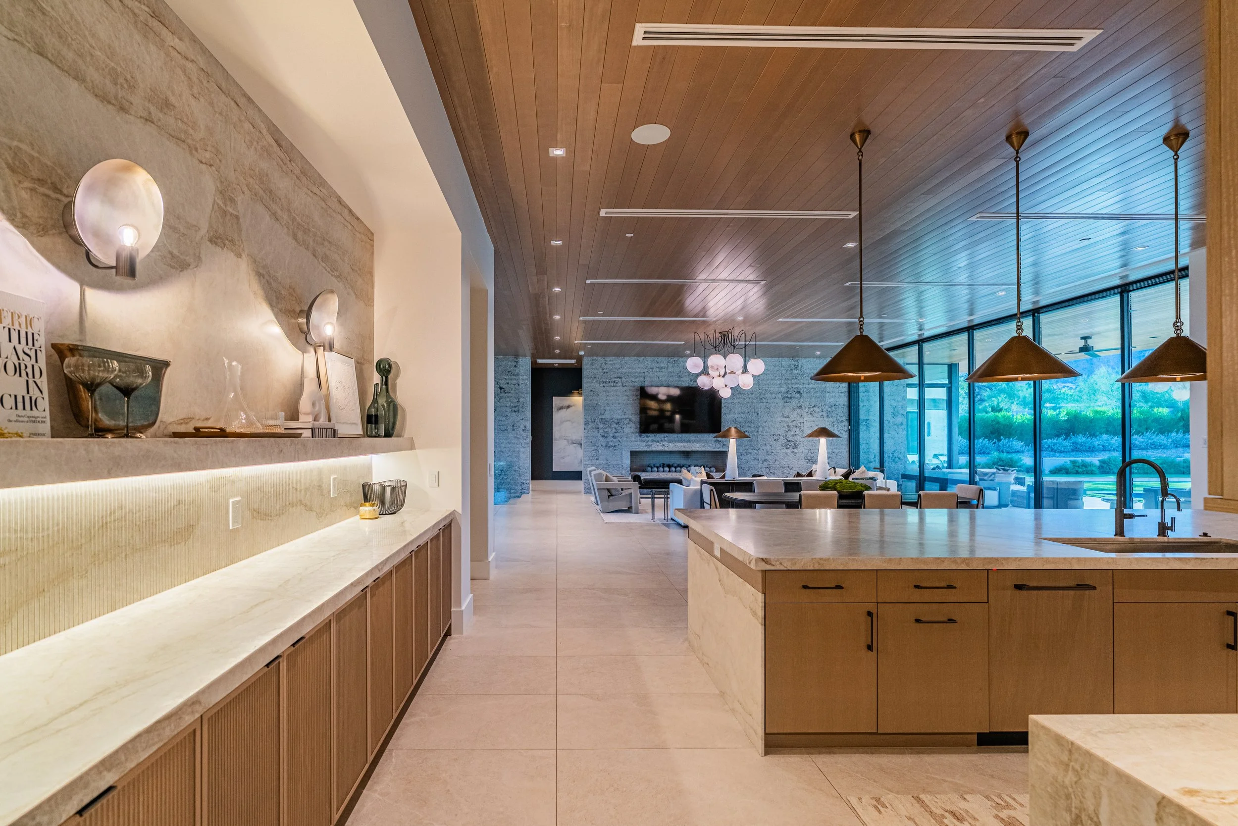 Modern kitchen with light stone countertops and wooden cabinetry, open to a living area with large windows and outdoor views.