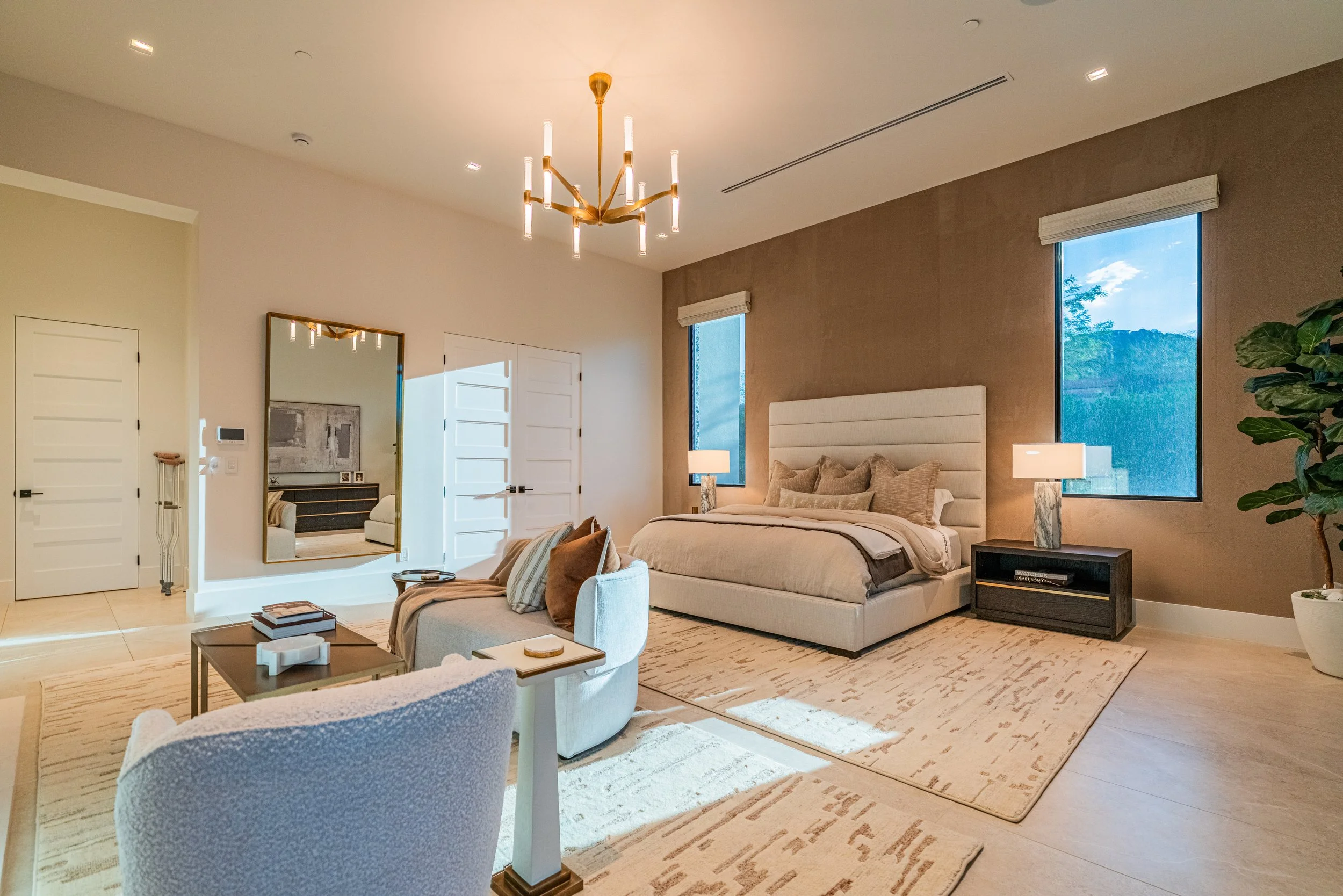 Modern bedroom with a large bed, side tables with lamps, a sitting area with chairs, a mirror, and large windows showing a blue sky with clouds and trees.