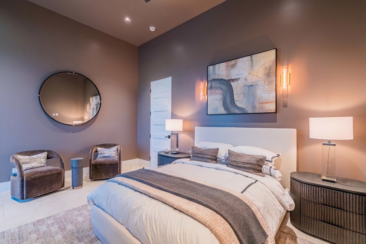 Modern bedroom with a white bed, abstract wall art, two bedside lamps, a mirror, and two armchairs in a neutral color.