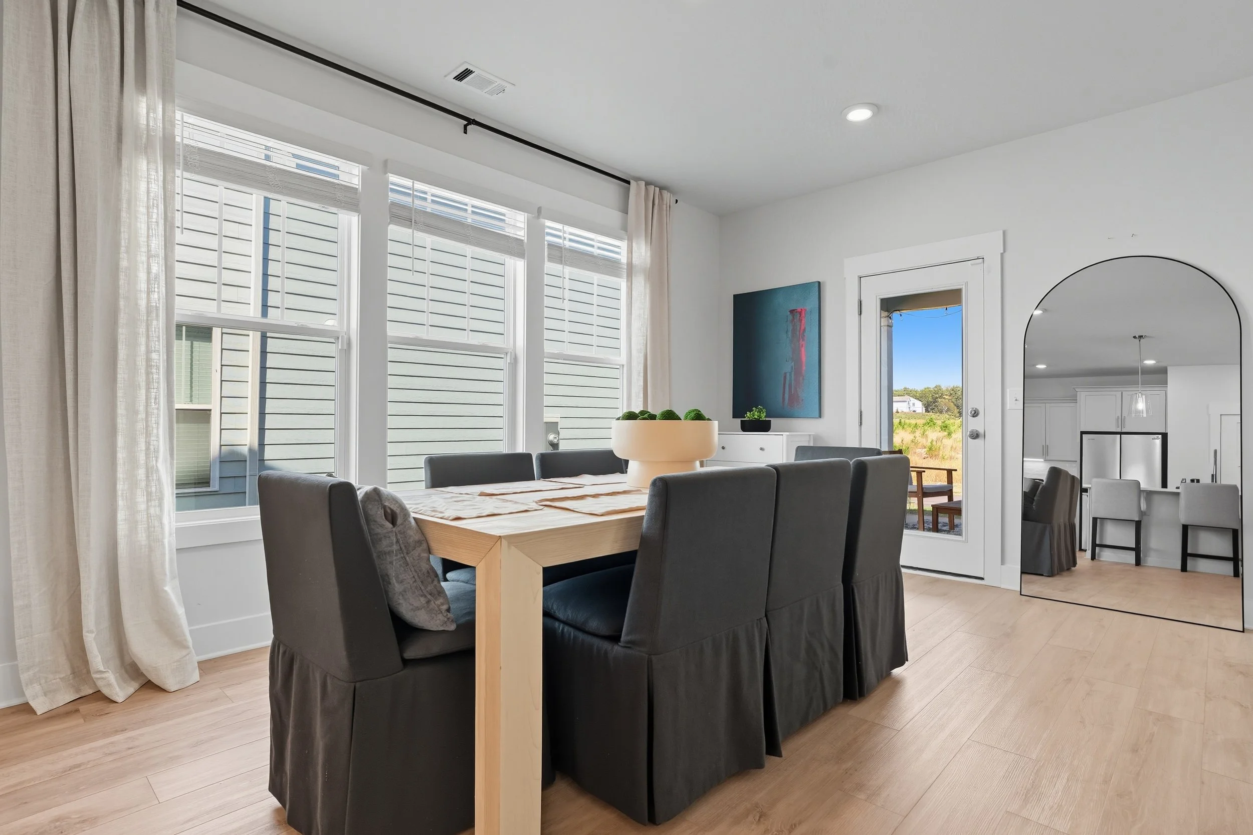 A dining area with a wooden table surrounded by six dark gray upholstered chairs, a large white planter with green decorative moss on the table, overlooking a bright room with large windows and an outside view of a grassy landscape and blue sky.