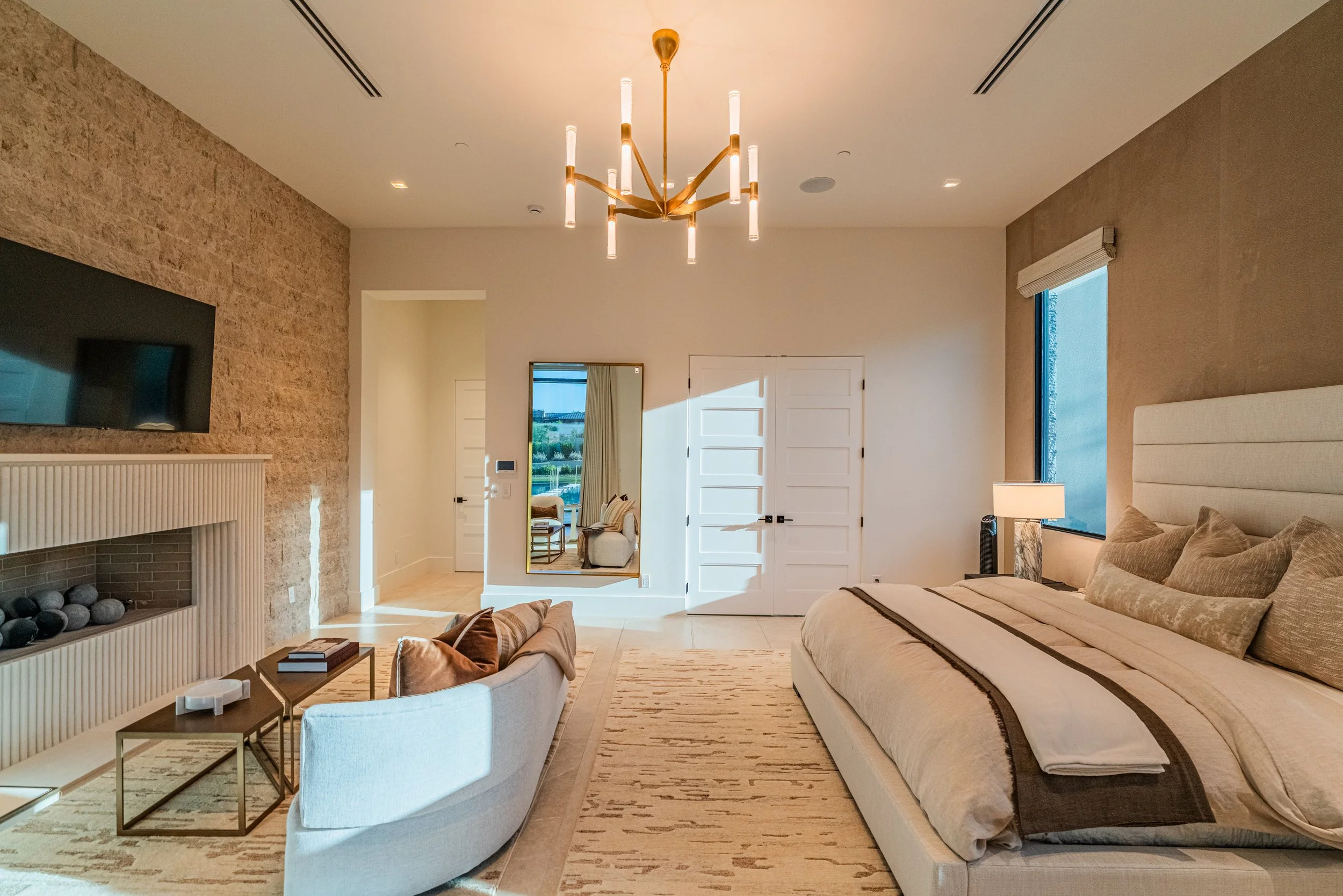 Living room with beige sofas, a cream armchair, a coffee table, a large mirror on the wall, a television above the fireplace, and a chandelier hanging from the ceiling. Bright natural light coming through a window on the right.