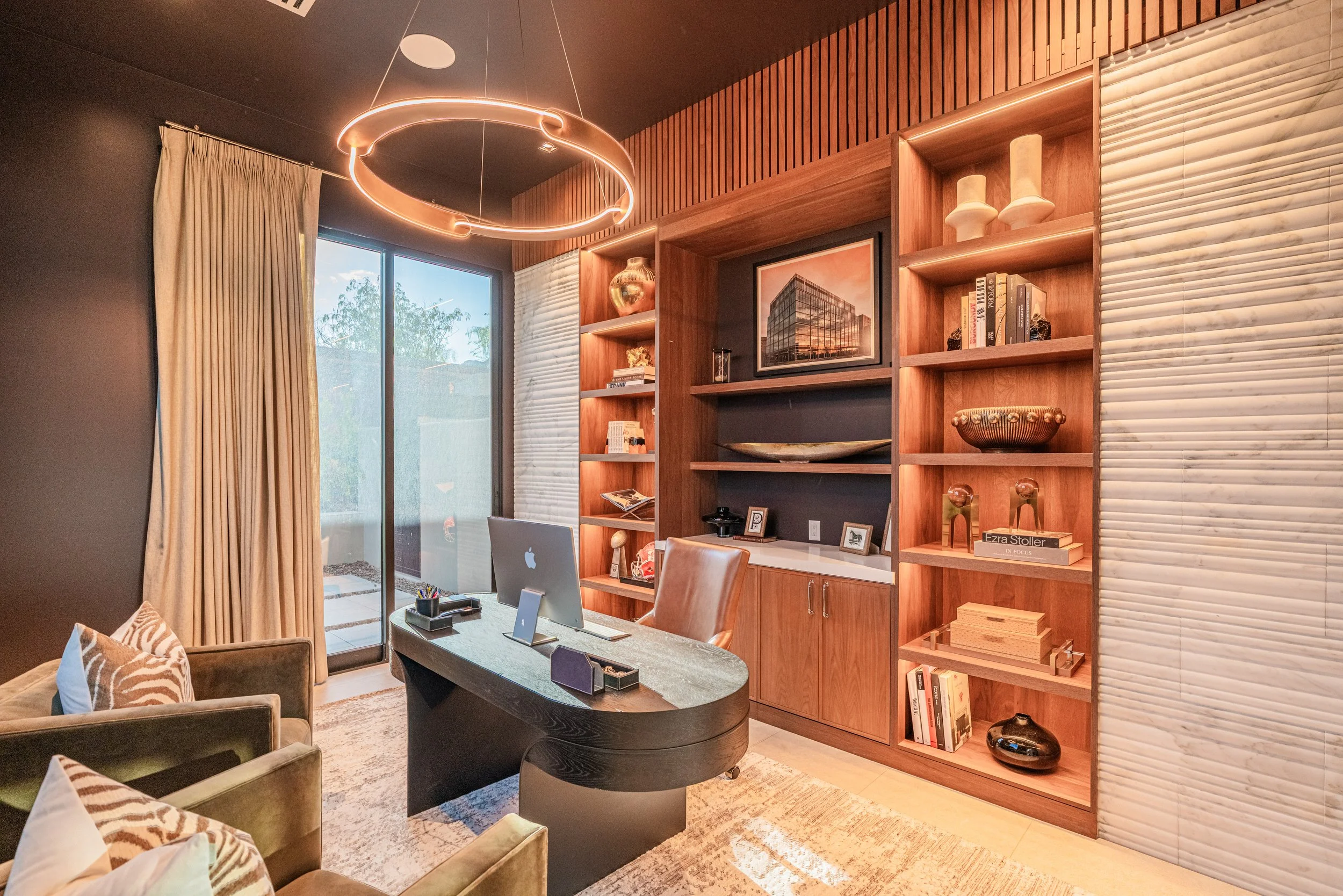A modern home office with a large wooden bookshelf, a desk with an iMac, beige chairs, a glass sliding door with beige curtains, and a circular ceiling light fixture.