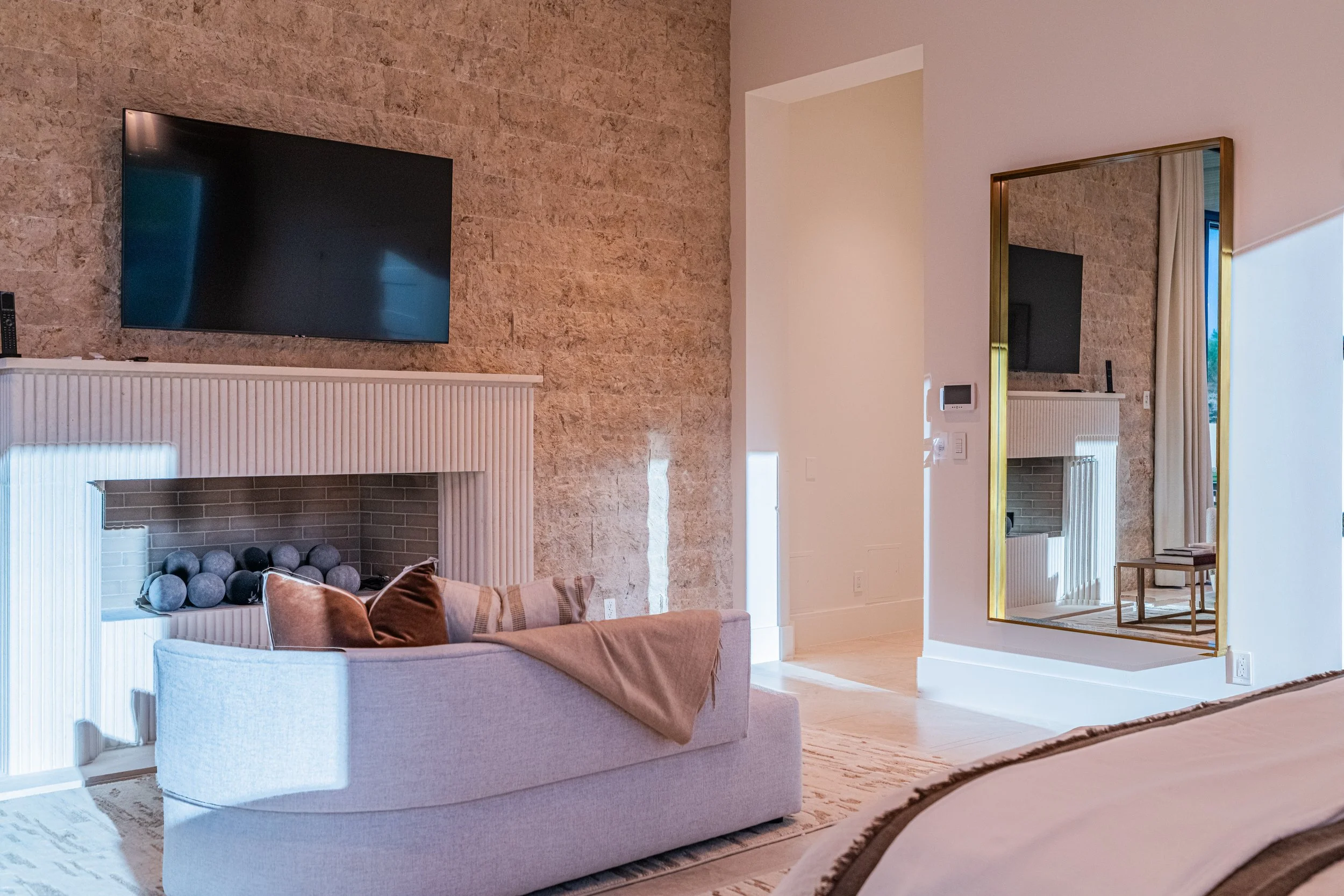 A living room with a beige sofa, a white fireplace with gray decorative balls inside, a wall-mounted flat-screen TV, a large mirror, and a window with curtains, featuring a modern interior design with natural light.