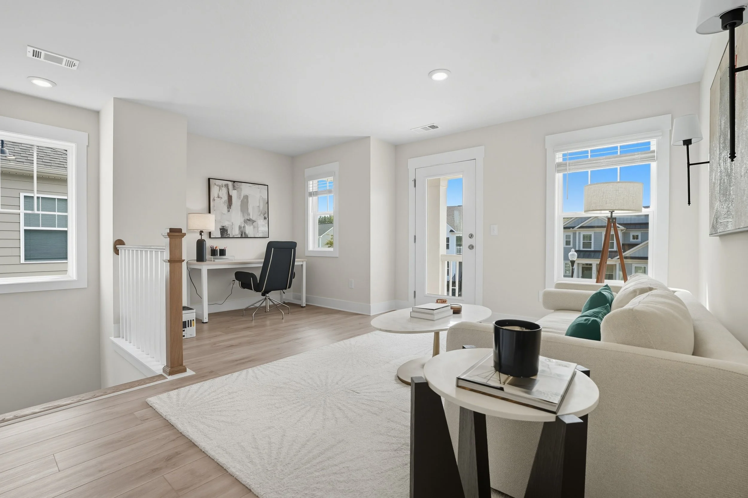 Bright living room with white walls, hardwood floors, a white sectional sofa with green pillows, a black end table with a book and a black mug, a white coffee table with a book, and large windows with white blinds letting in natural light.