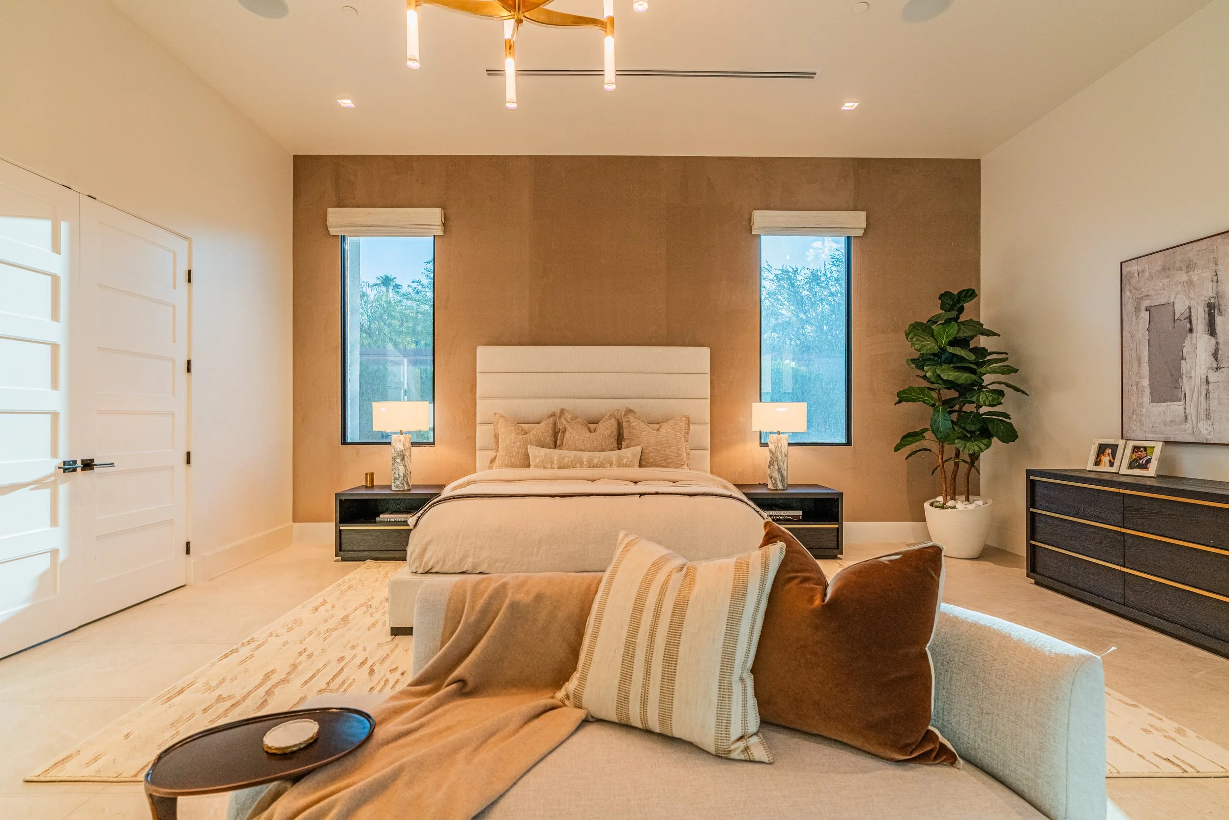 A bedroom with a large bed featuring a high, upholstered headboard, two bedside lamps, two windows behind the bed, a dresser with picture frames, a potted plant, and a gray sofa with a striped pillow, viewed from a person's legs resting on it.