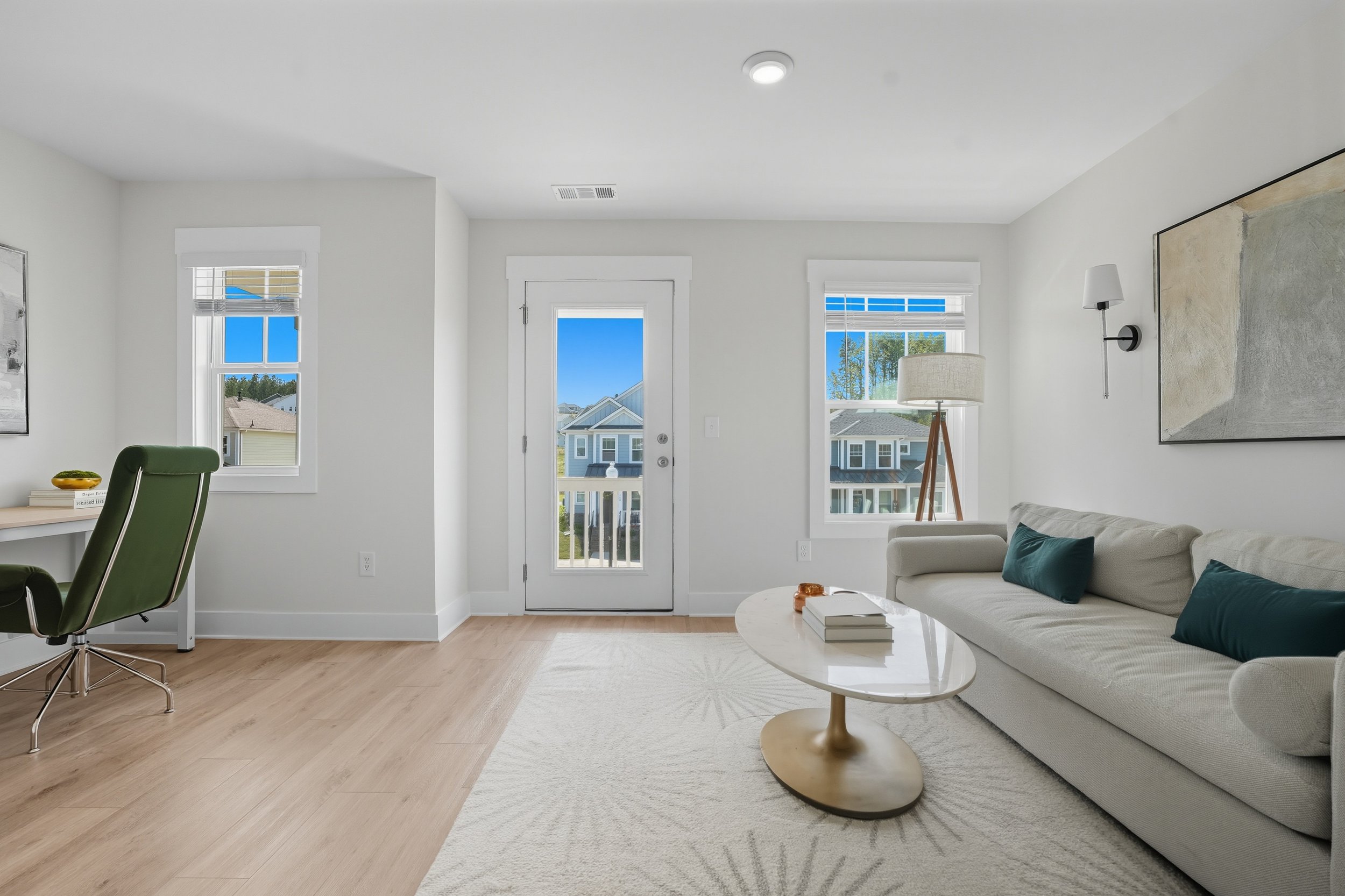 Bright living room with white walls, three windows with white blinds, a gray sofa with blue cushions, a round coffee table with books and a tray, a floor lamp, and artwork on the wall, with a view of neighboring houses outside.