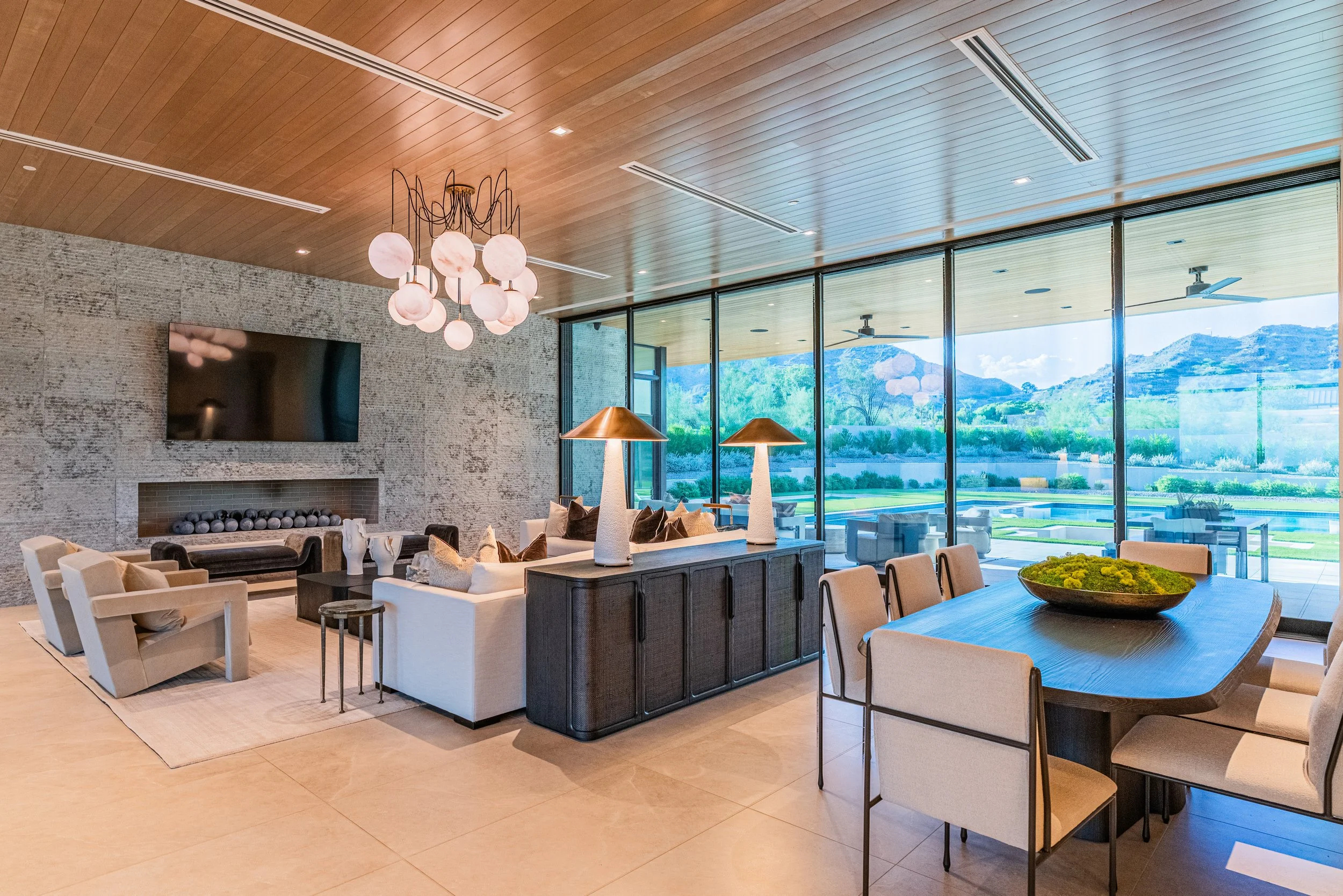 Modern living room with large glass windows showing a scenic outdoor landscape, featuring a chandelier, a fireplace, contemporary furniture, and a wooden ceiling.