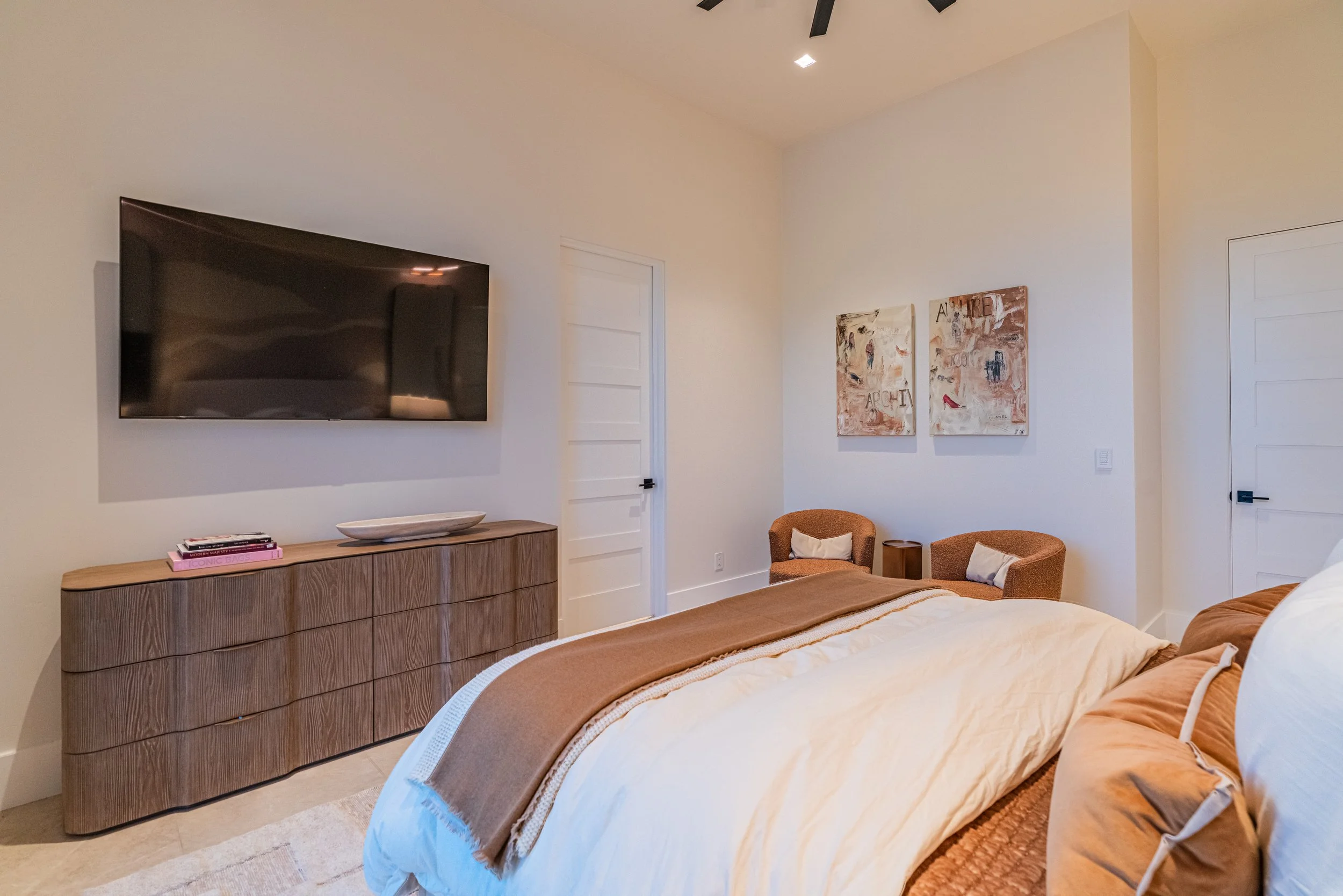 A bedroom with a wooden dresser, wall-mounted TV, two armchairs, artwork on the wall, and a bed with beige and white bedding.