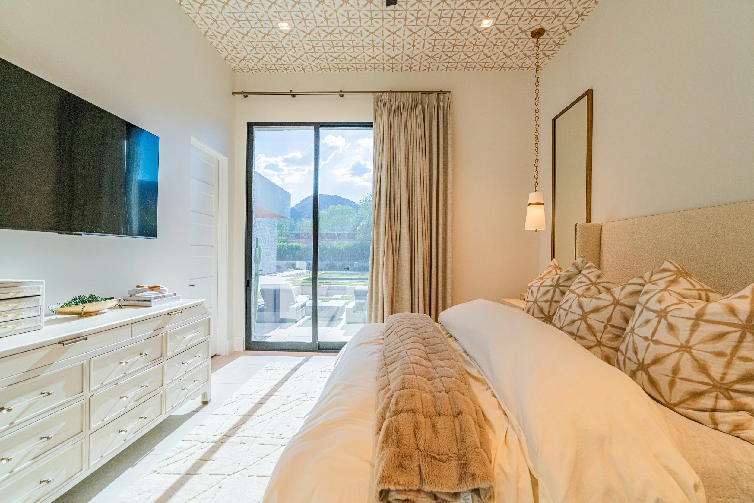 A bedroom with a large bed adorned with pillows, a dresser with decorative items, a wall-mounted TV, a sliding glass door with curtains leading to an outdoor patio, mountain views, a patterned ceiling, and warm lighting.
