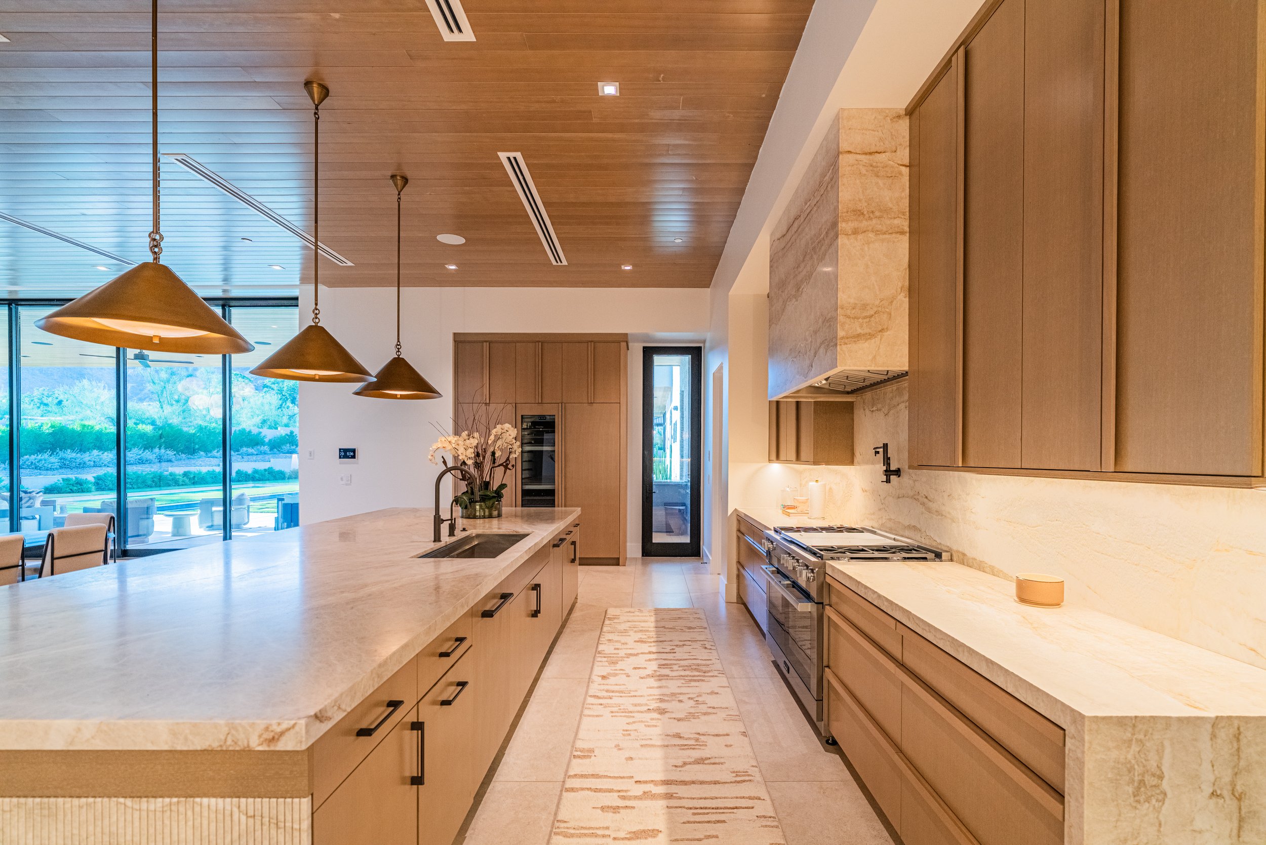 Modern kitchen with beige cabinets, marble countertops, a gas stove, pendant lights, a vase with white flowers, and large windows showing outdoor scenery.