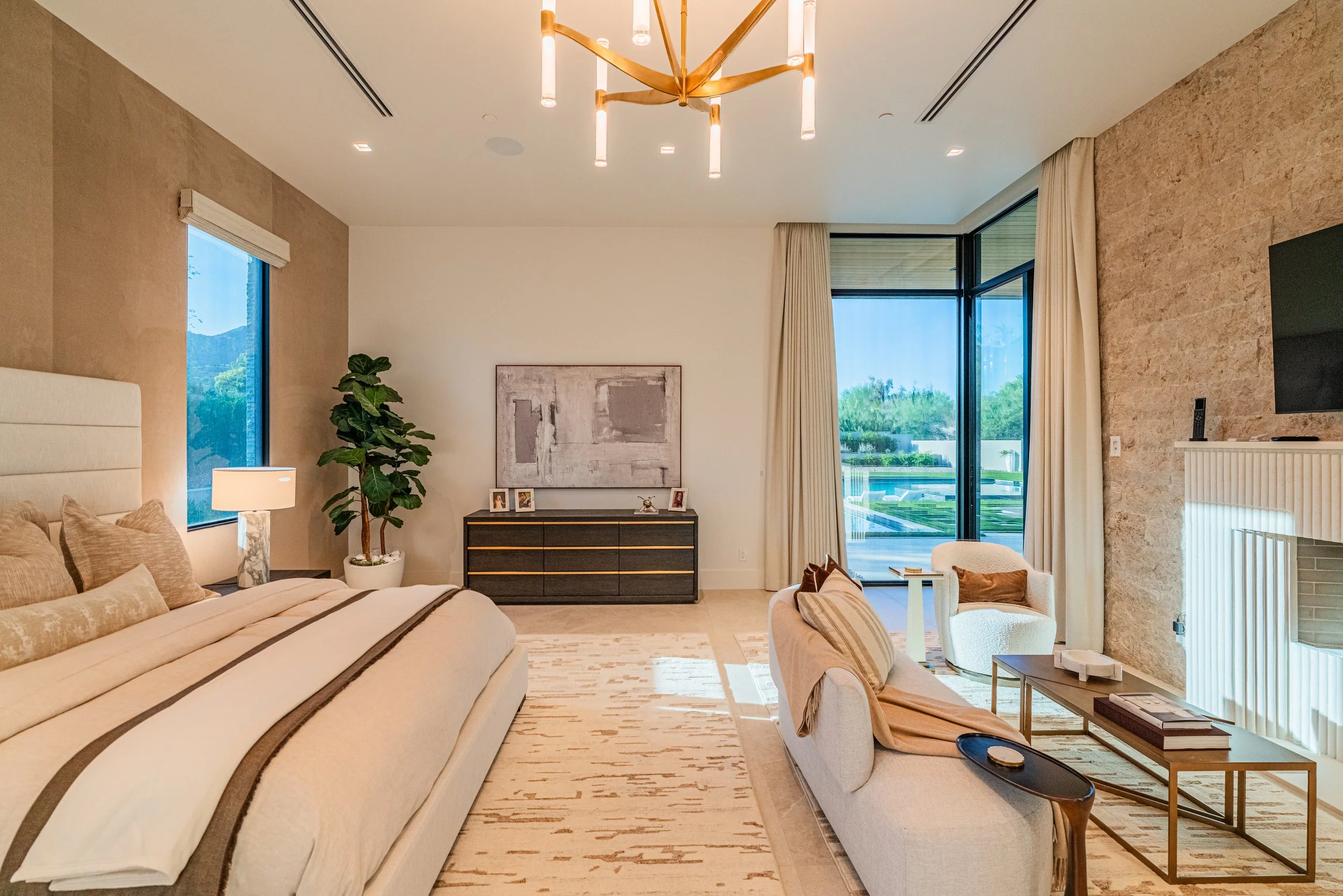 Living room with large windows, beige and cream furniture, modern chandelier, a plant, and decorative artwork on the wall.