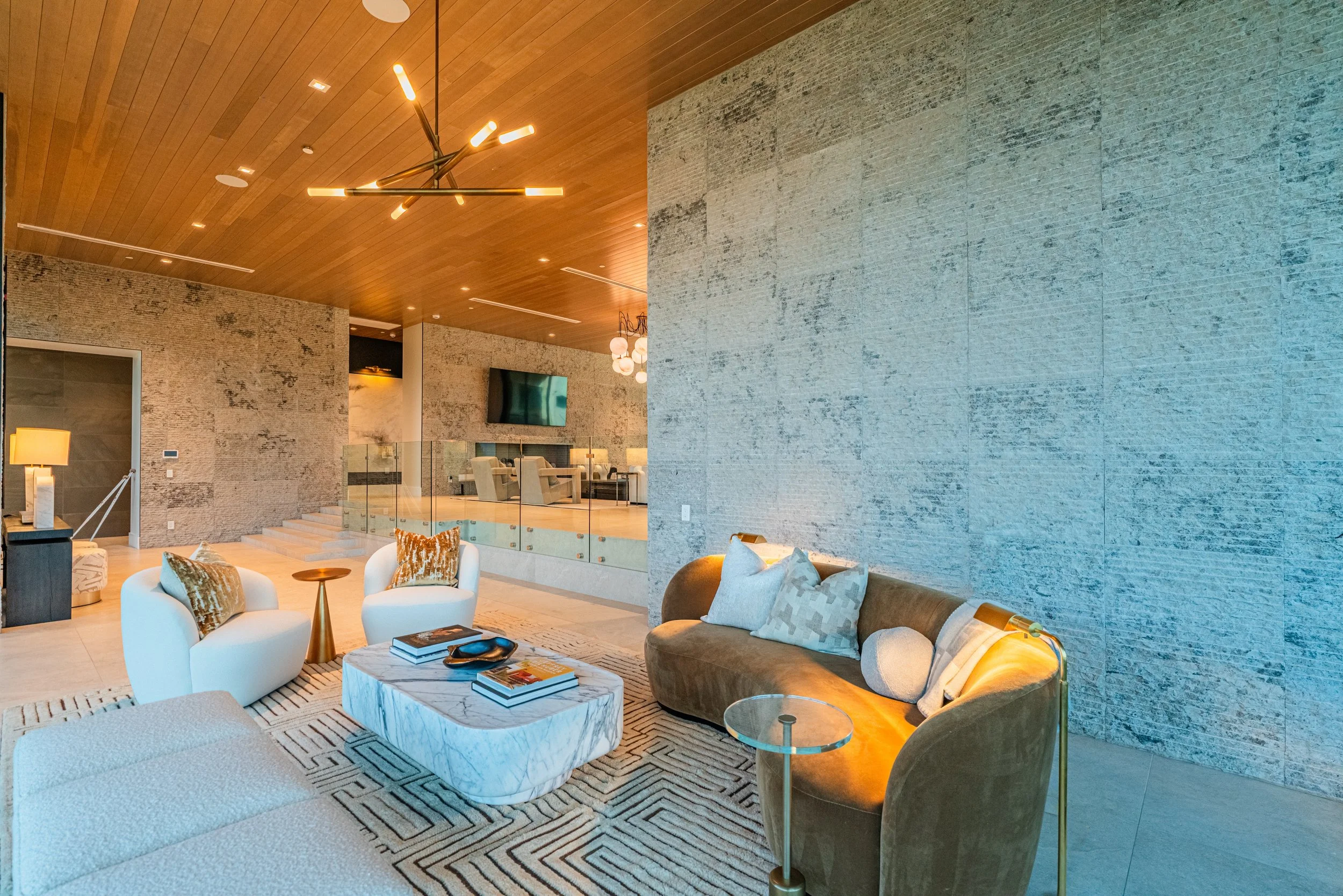 Modern living room with wooden ceiling, stone accent walls, contemporary lighting fixtures, white and beige furniture, and decorative pillows.