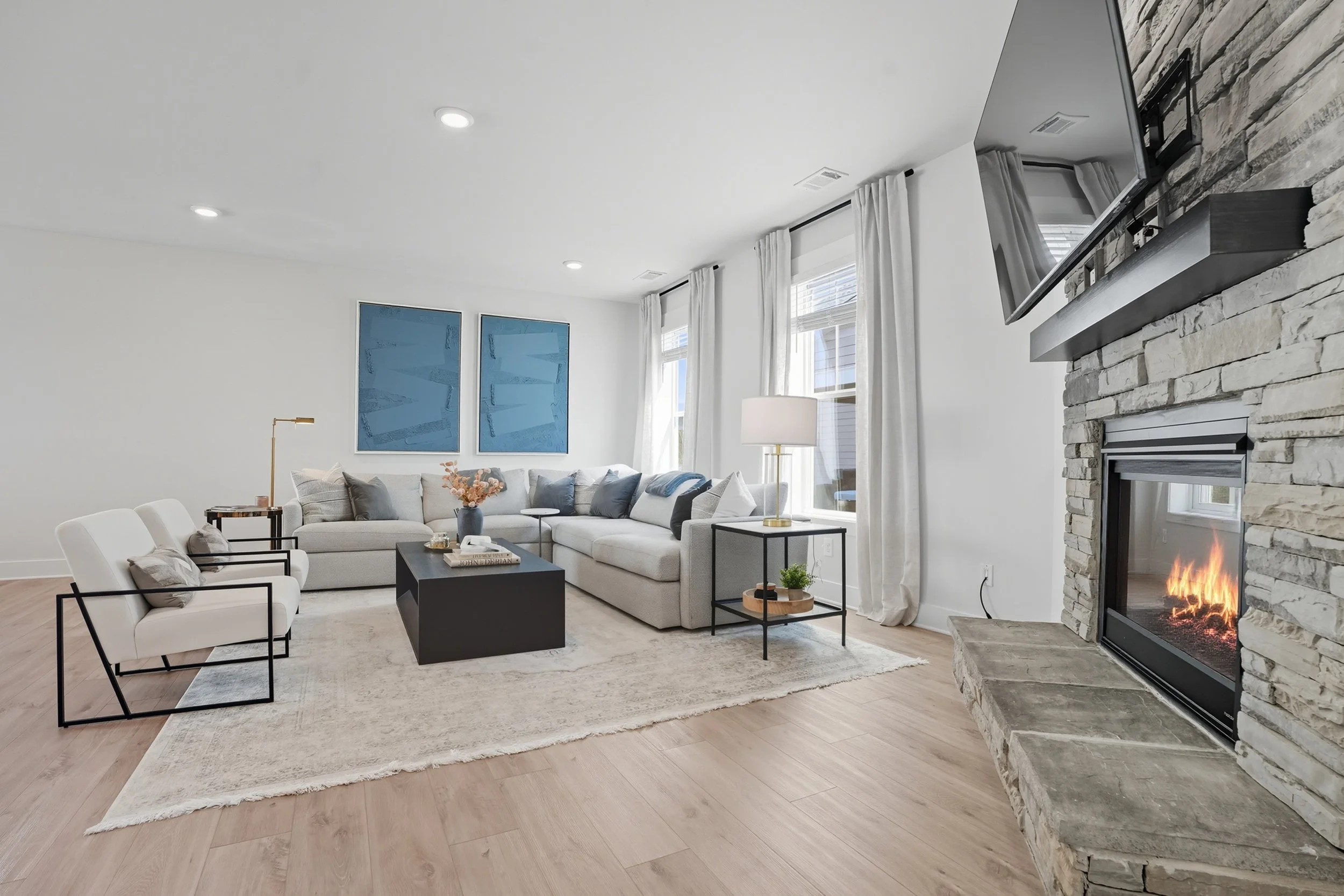 A modern living room with a light-colored sectional sofa, black and white chairs, a black coffee table, and a fireplace with a stone surround. There are three large windows with white curtains and blue artwork on the wall. A floor lamp and a small pl