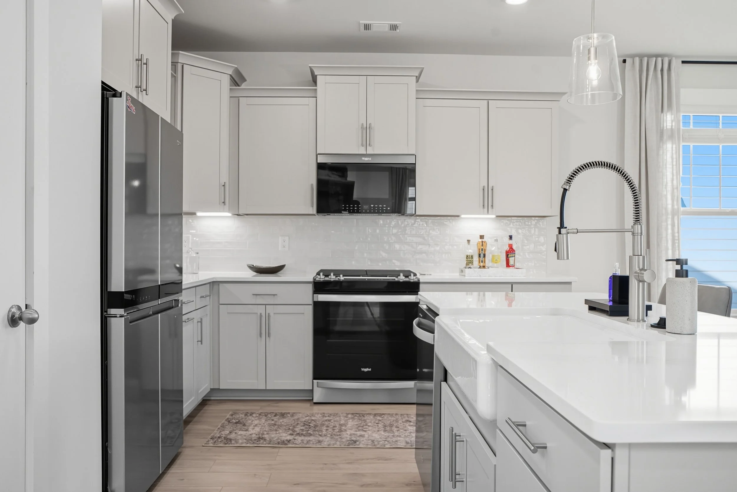 Modern white kitchen with gray cabinets, stainless steel appliances including a refrigerator, oven, and microwave, and a white island with a sink and a gooseneck faucet. Bright natural light through a window with white curtains.
