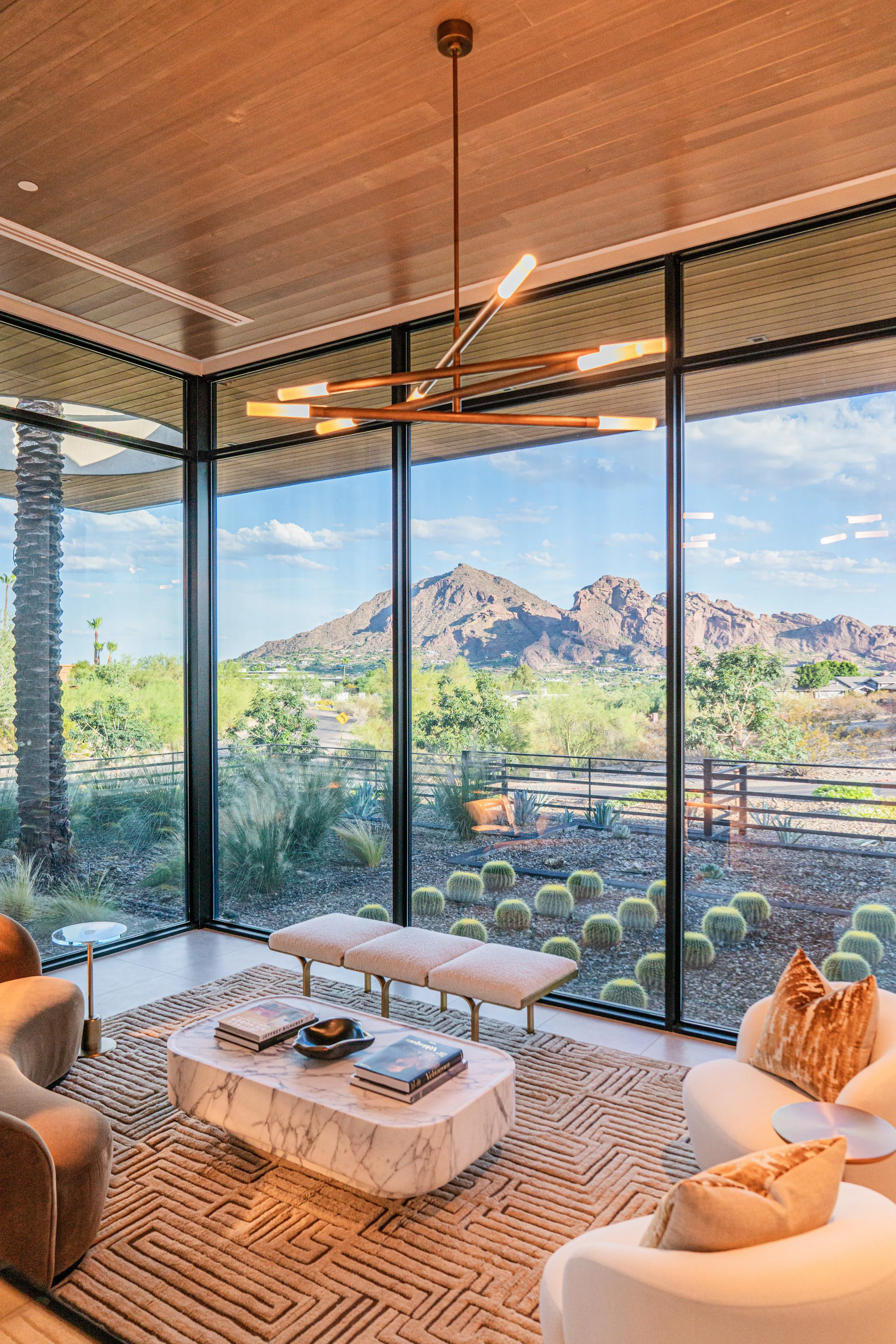 Modern living room with large floor-to-ceiling windows showing desert mountains and blue sky outside. The room has a marble coffee table with books, a patterned rug, and beige and white seating with pillows, and a unique overhead light fixture.