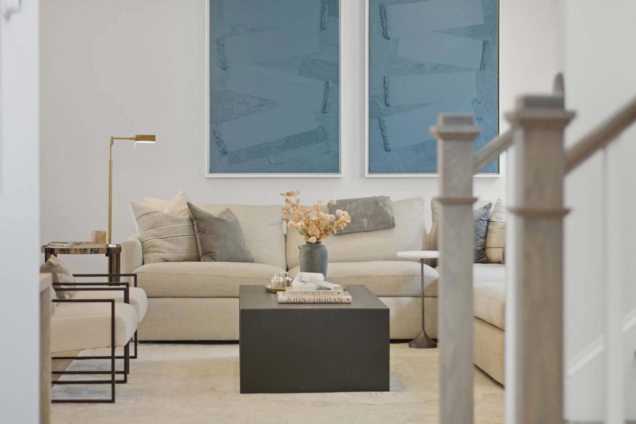 Living room with beige sofa, dark coffee table with books and decor, gold floor lamp, abstract blue artwork on white wall, beige armchair, floral vase.