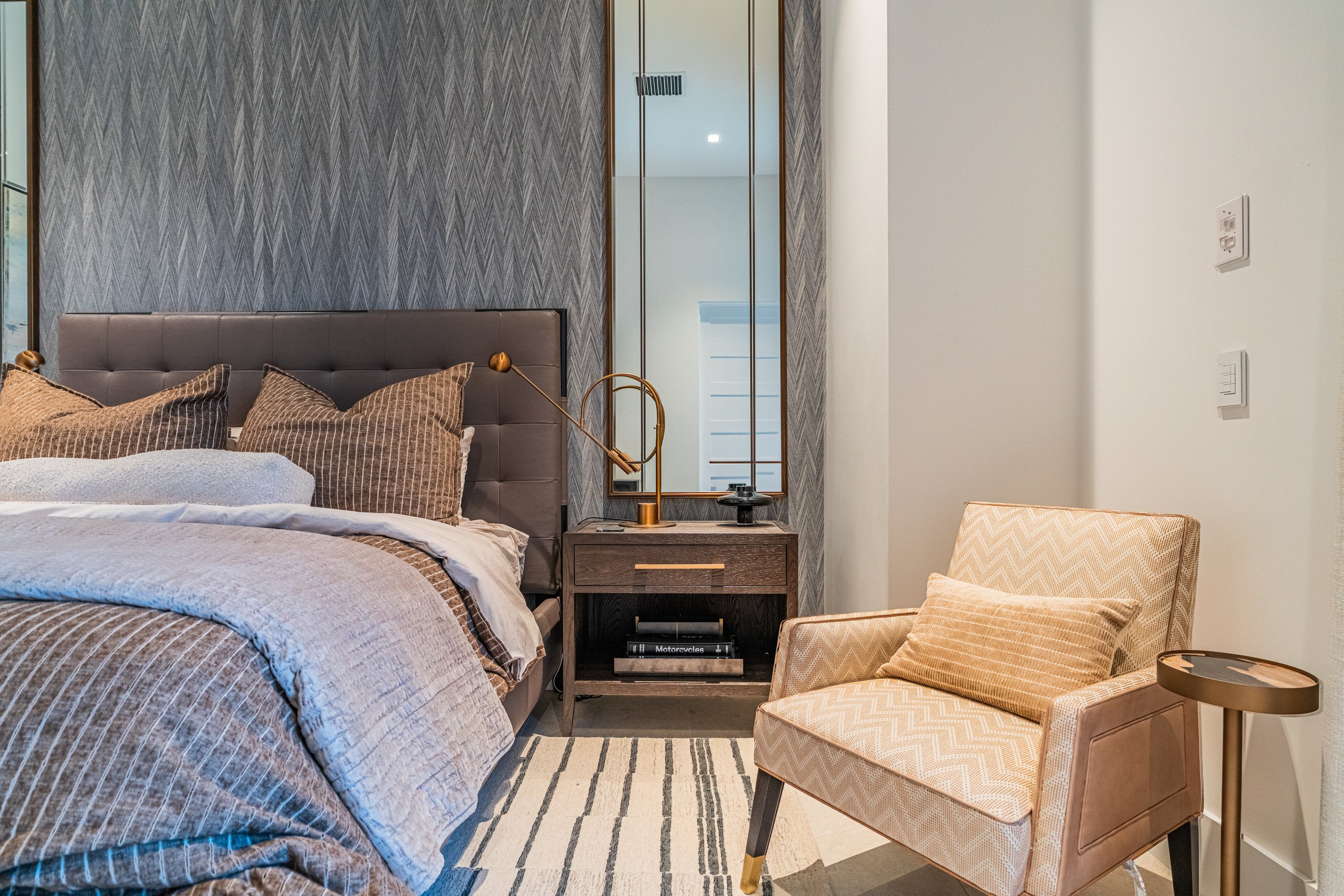 A bedroom with a bed featuring a grey upholstered headboard and striped bedding, a nightstand with a modern gold and black adjustable lamp, a beige patterned armchair, and a large mirror on the wall reflecting a staircase and white wall with switches