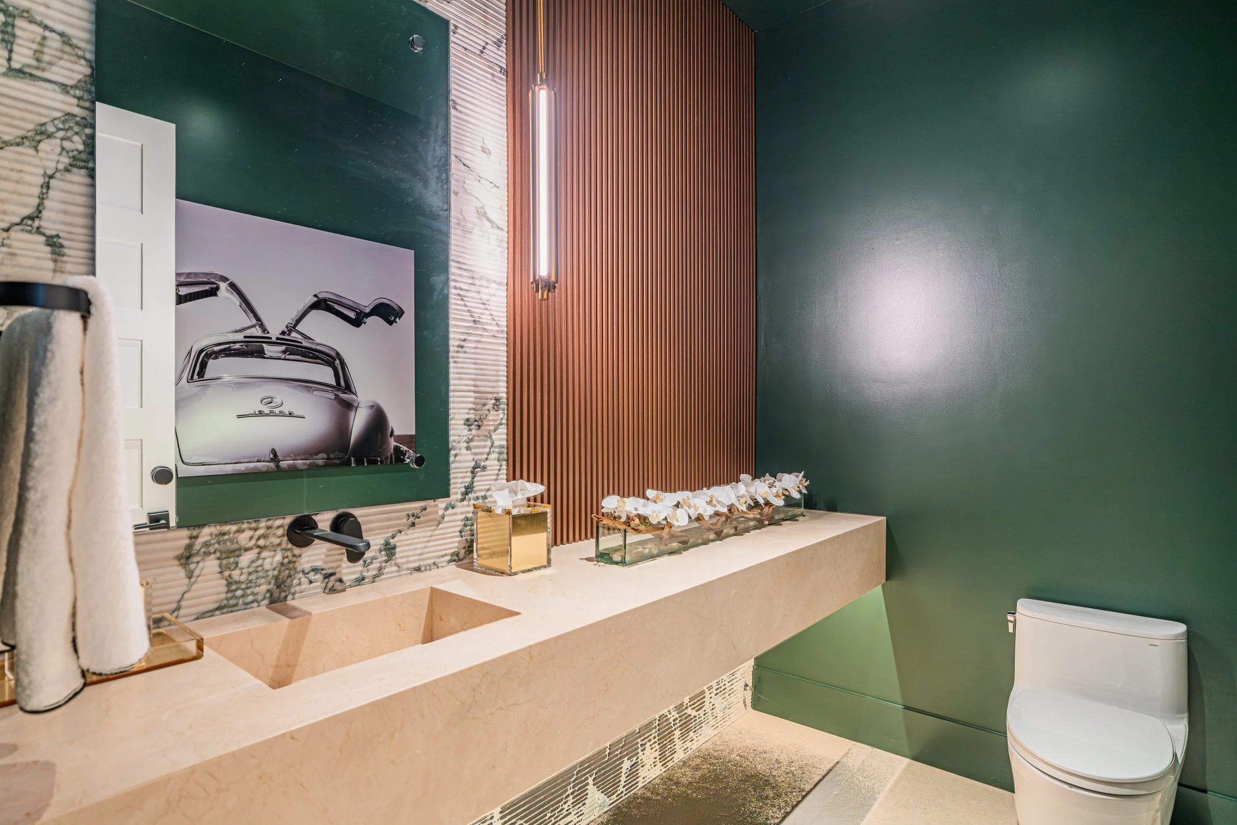 Modern bathroom with green walls, a beige marble countertop with a built-in sink, a large mirror, a hanging vertical light fixture, decorative flowers, and a white toilet.