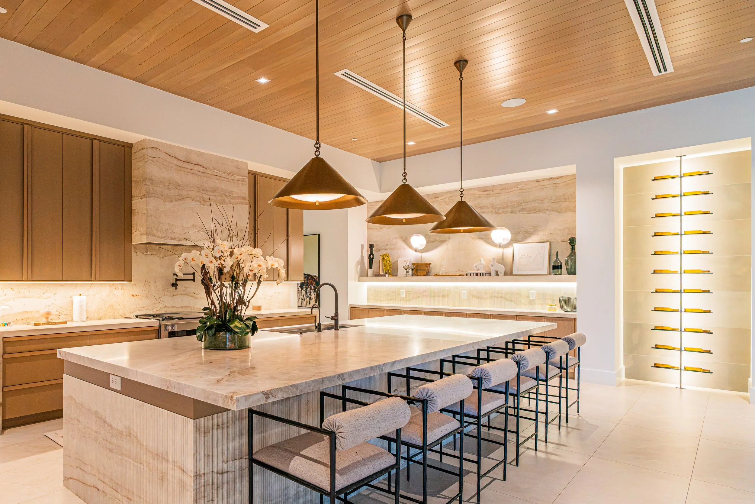 Modern kitchen with a large marble island, pendant lights, wooden ceiling, and built-in shelves with yellow accents.