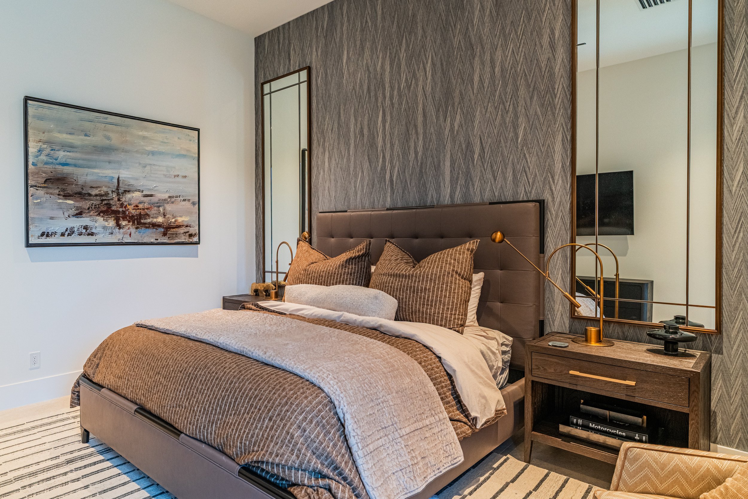 A modern bedroom with a bed, pillows, two nightstands, a large abstract painting on the wall, a decorative mirror, and built-in closet with mirrored sliding doors.