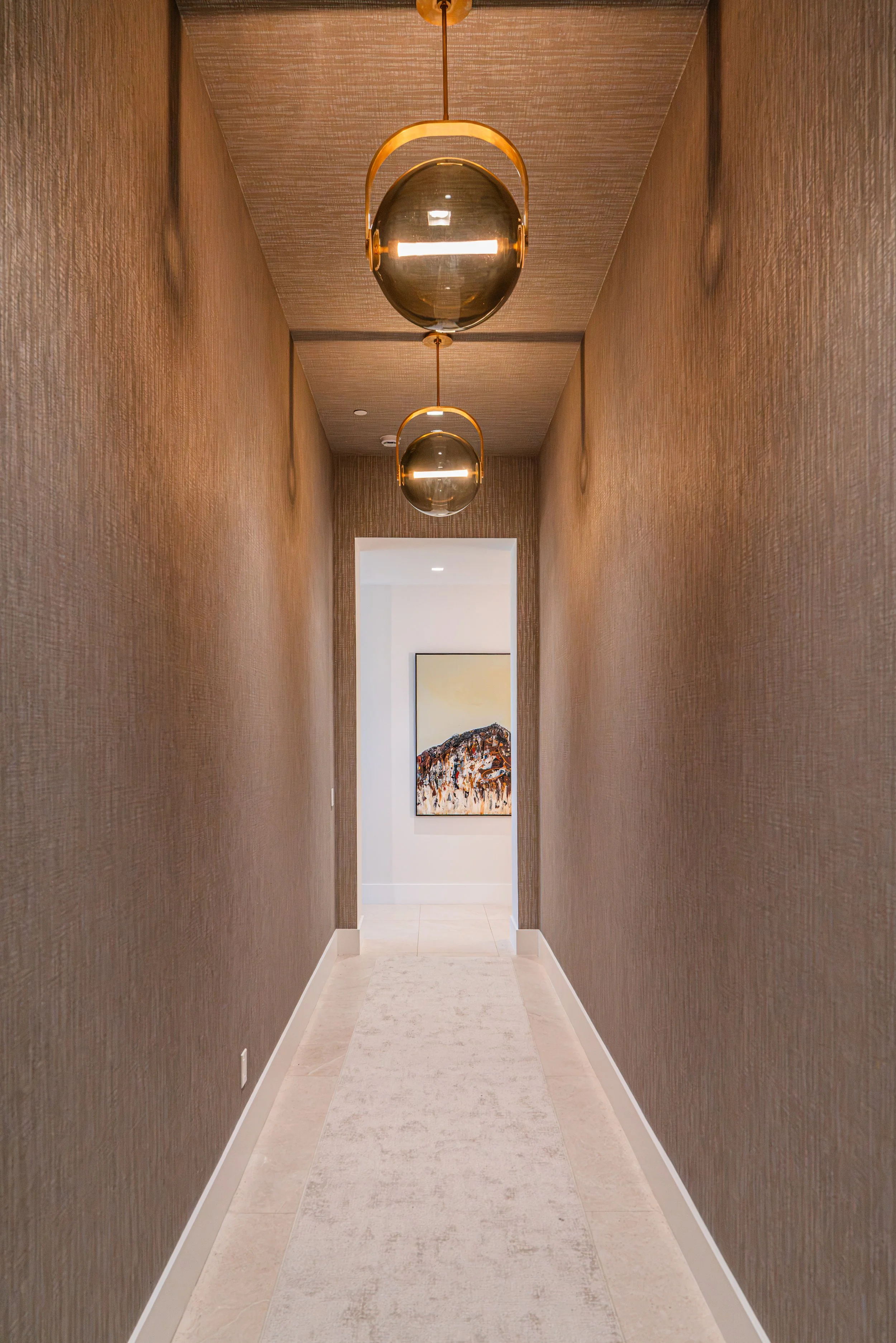 A narrow hallway with textured brown walls, a light-colored tiled floor, and modern pendant lights hanging from the ceiling. At the end of the hallway, an opening reveals a room with a framed painting on the wall.