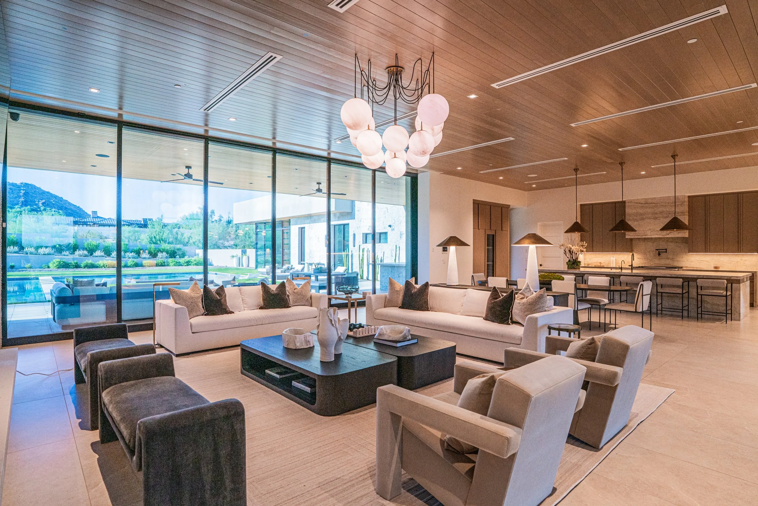 Modern living room with beige sofas, black and gray chairs, a wooden coffee table, and large windows showing an outdoor patio, pool, and greenery. Ceiling features wooden panels and a large chandelier with white globes.