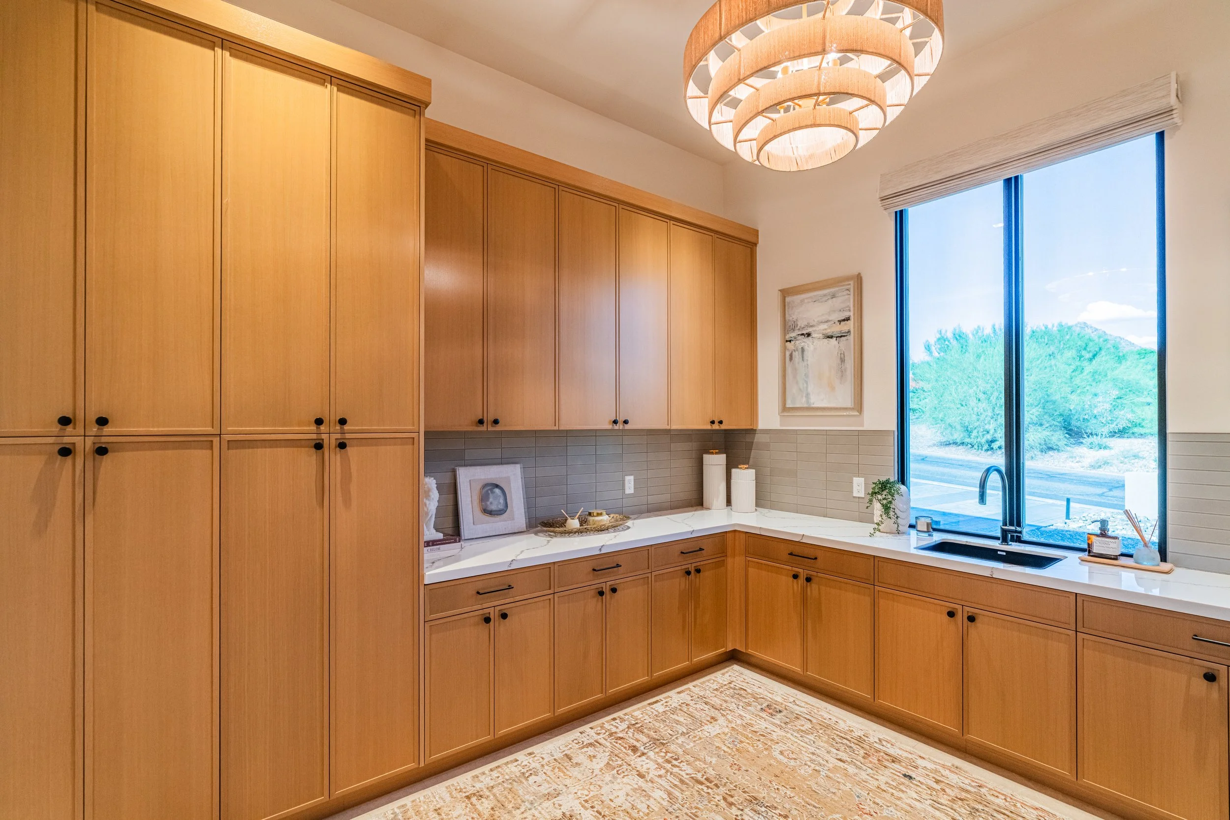 Modern kitchen with wooden cabinetry, white countertop, large window with a view of green trees, and a contemporary chandelier overhead.