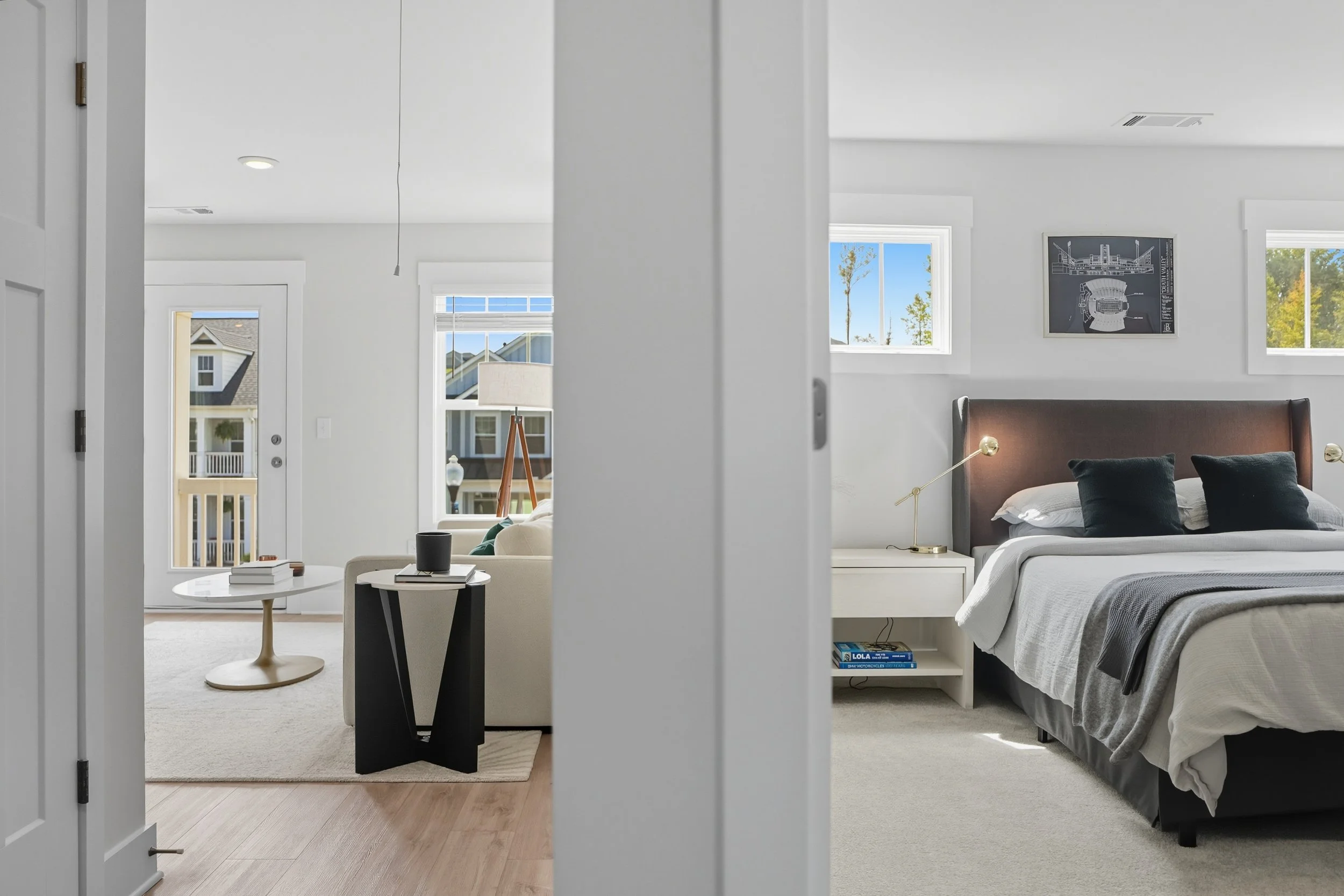 View of a modern bedroom with white walls, a bed with gray and black pillows, and a white nightstand. There is a large window showing trees outside. Adjacent is a living room with a white ceiling, a small round table, chairs, and a door leading outsi