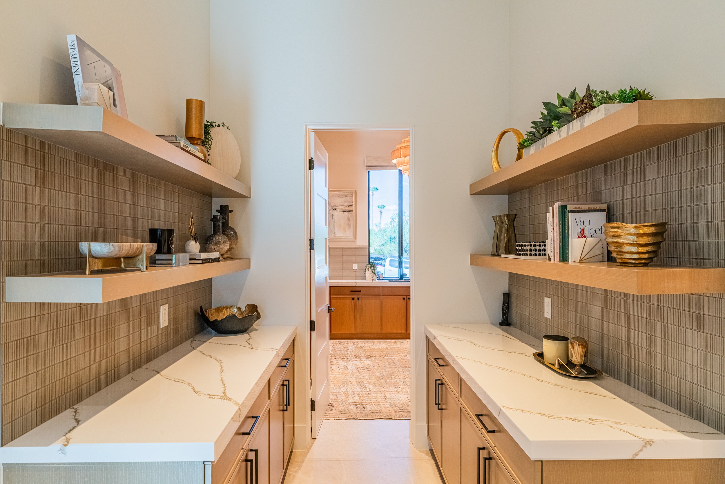 A modern kitchen with light wood cabinetry, white marble countertops with gold veining, open wooden floating shelves decorated with vases, books, and greenery, and a view into an adjacent room with a sliding glass door and outdoor scenery.