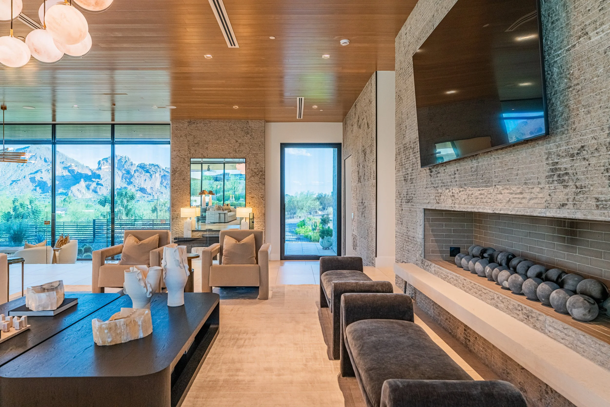 Interior of a modern living room with large windows showing mountain scenery, light wood ceiling, stone and brick walls, a fireplace with black stones, and furniture including beige armchairs, a dark wood table with decorative objects, and a wall-mou
