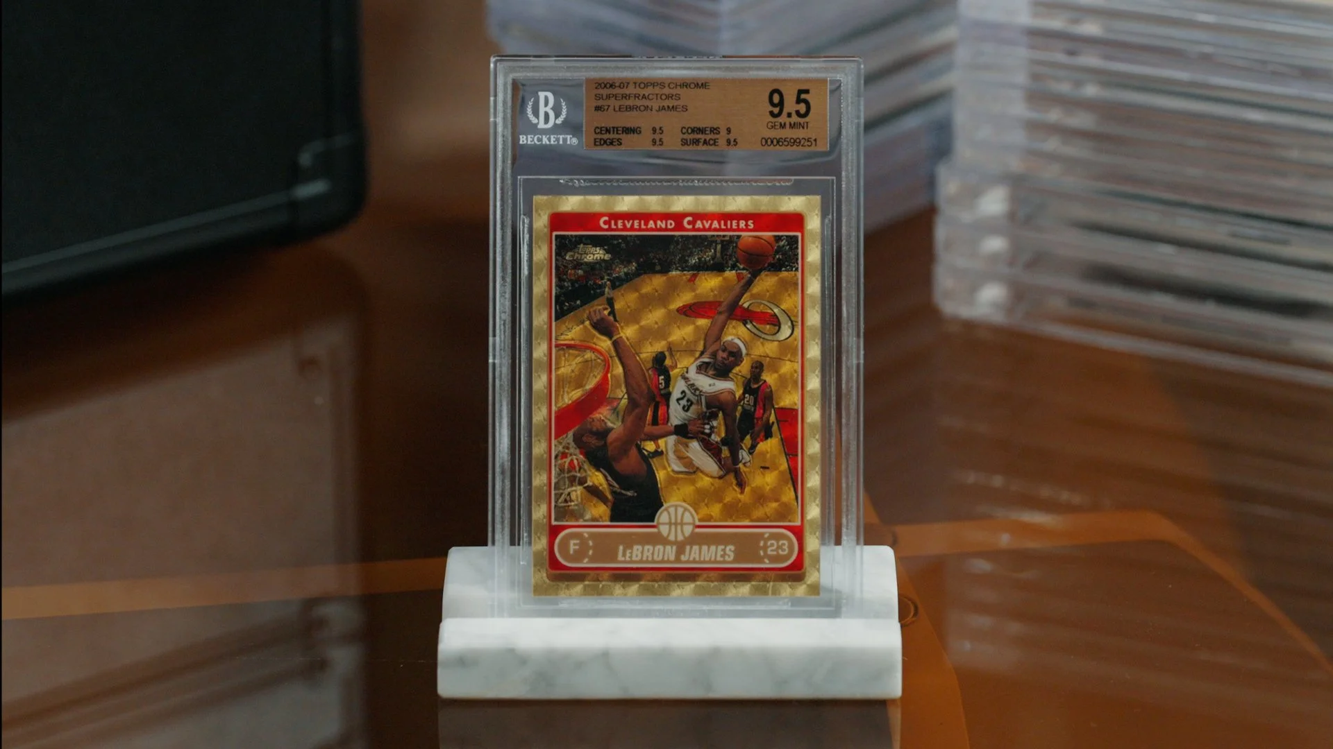 A LeBron James basketball trading card encased in a clear plastic case, displayed on a white stand. The card features LeBron James in a Cleveland Cavaliers uniform, jumping with a basketball. The background of the card is gold with a holographic pattern.
