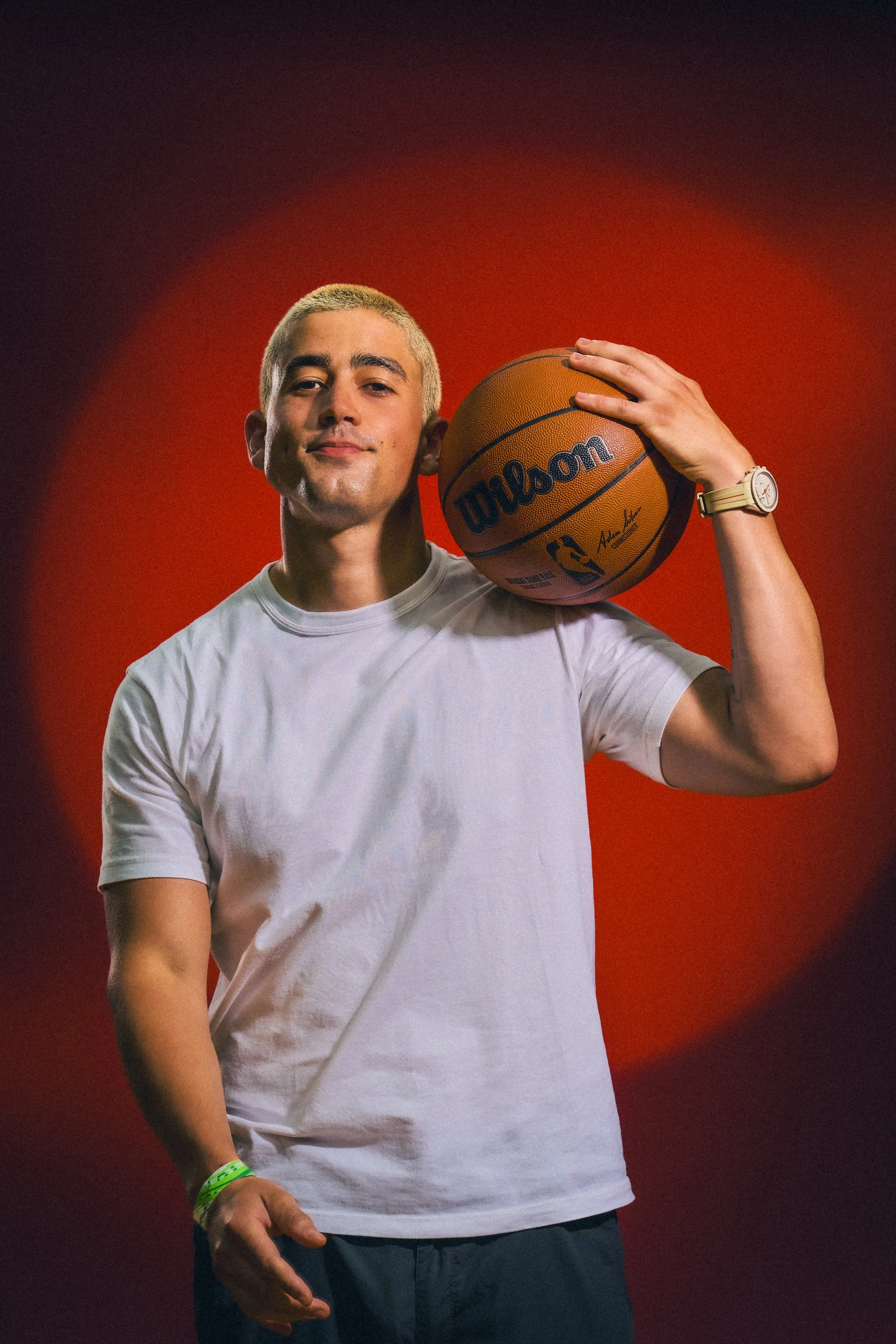A man with blonde hair, wearing a white t-shirt, holding a basketball on his shoulder, standing against a dark red background.