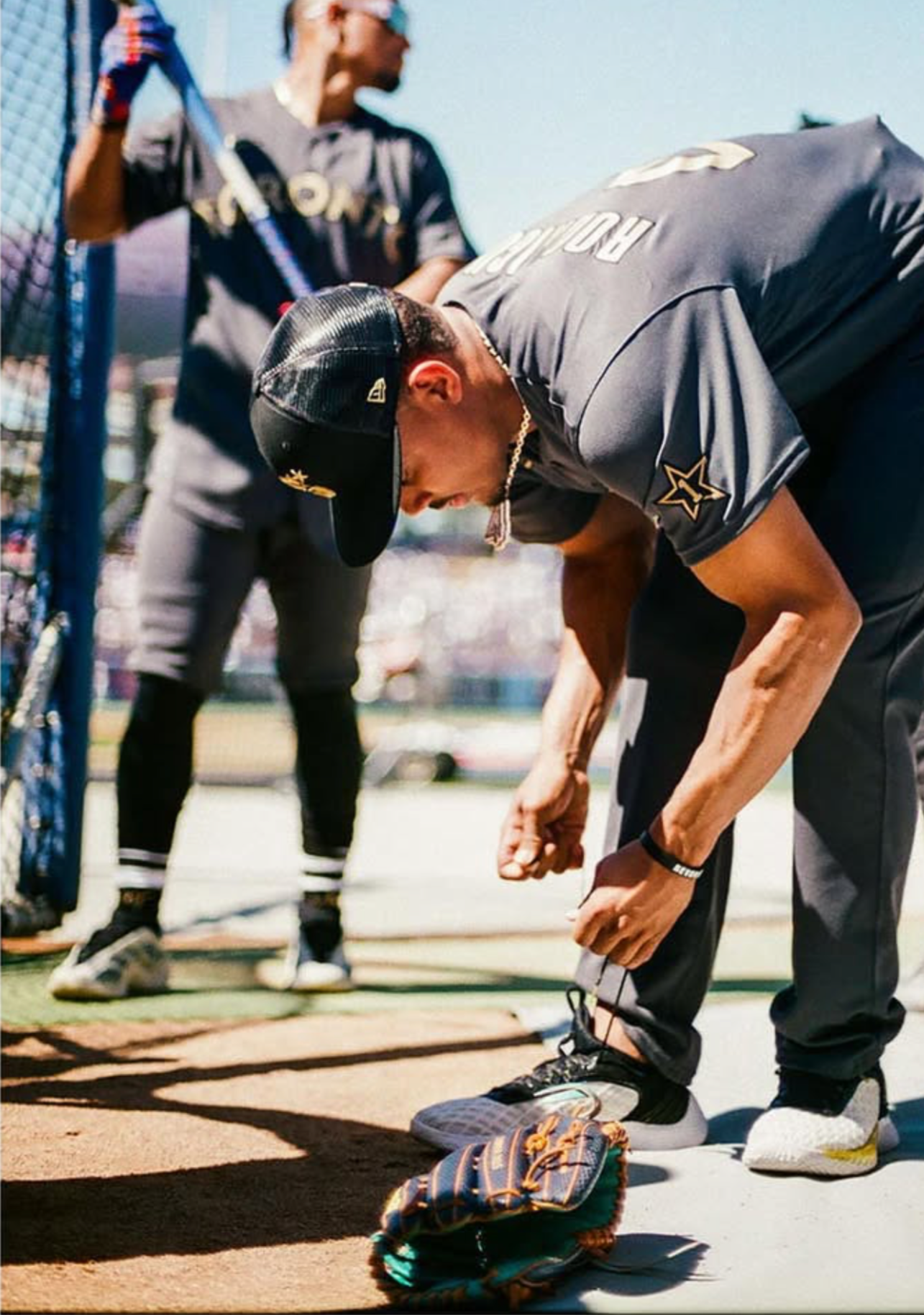 Two young men at a baseball field, one is bending down tying his shoe, wearing a black cap and gray athletic clothing, while the other stands in the background holding a bat, dressed in sports gear, near a blue baseball fence.