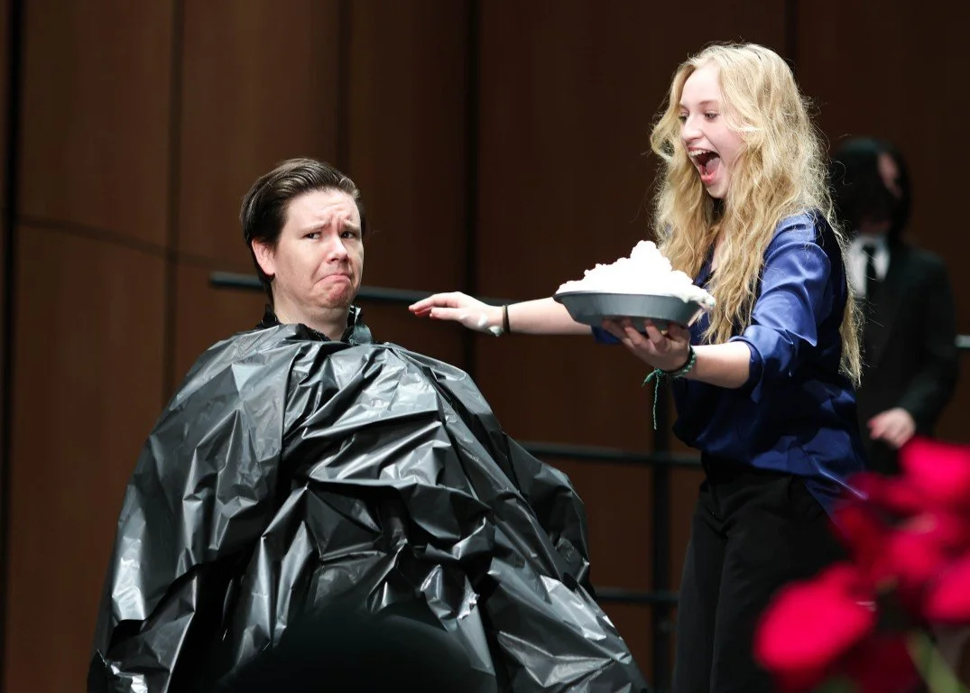 A woman with long blonde curly hair offering a bowl of whipped cream or snow to a man with short dark hair who is seated and wearing a black plastic cape, in a room with wooden walls.