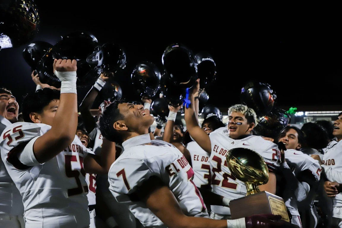 High school football team in white uniforms celebrating victory, lifting helmets and holding a trophy after winning a game.