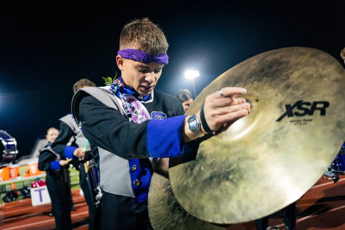 A young performer in a marching band uniform and purple headband adjusting a large brass cymbal on a football field at night.