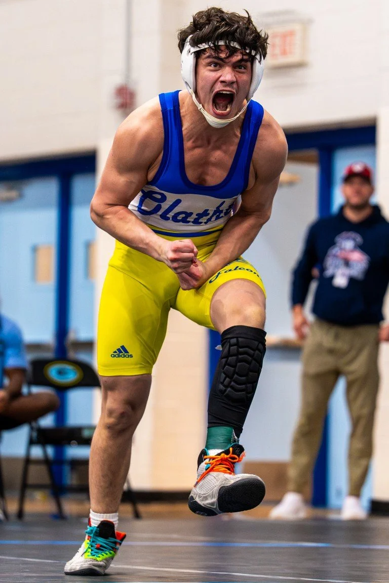 A male athlete in a blue and yellow uniform celebrating with a clenched fist and an open mouth, wearing a protective knee brace, headphones, and colorful running shoes, in an indoor sports facility.