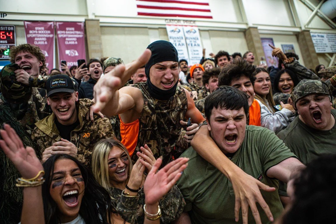 A group of young people wearing camouflage and casual clothing celebrating and cheering at an indoor event, with some making expressive gestures and faces.