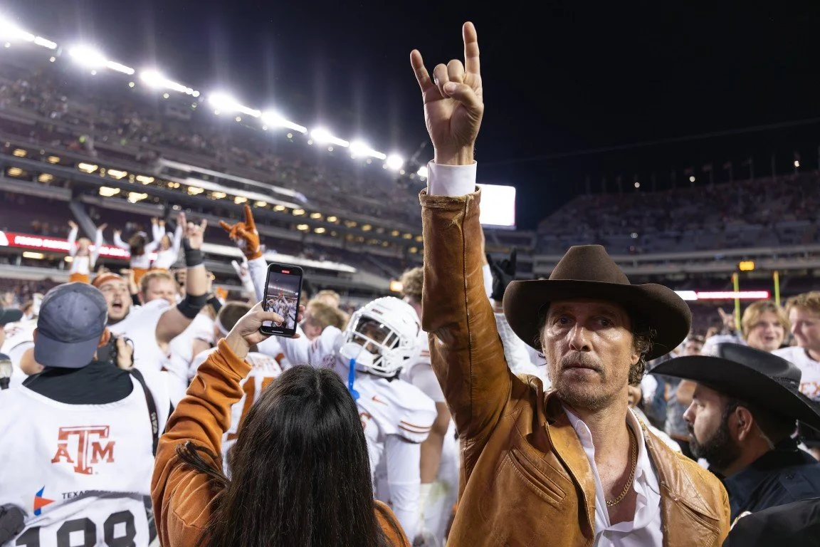 Person dressed as Indiana Jones at a night football game, surrounded by players and fans, holding up a hand in a peace sign.