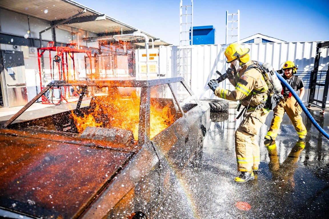 Firefighters in full gear spray water on a burned, rusted vehicle on a wet surface, with metal structures and a white storage container in the background.