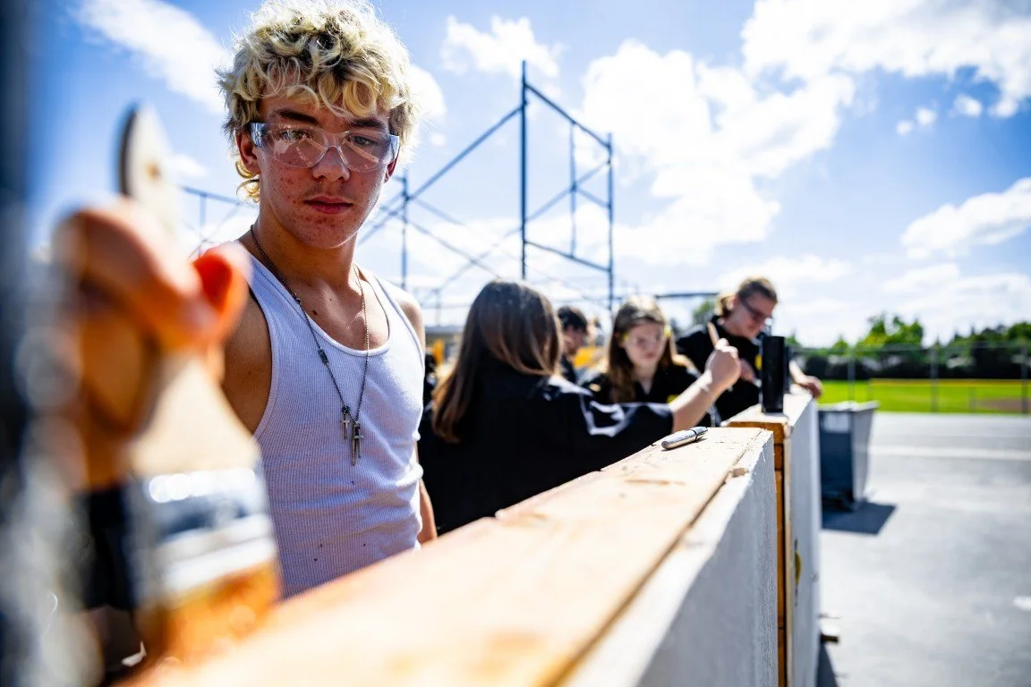 A young man with blond curly hair and safety goggles, holding a paintbrush, standing at a construction site outdoors, with other people working in the background.