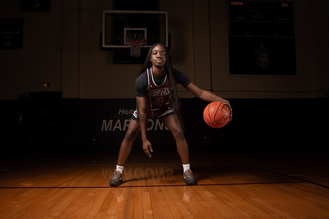A female basketball player in a dark indoor gym, holding a basketball, in a defensive stance, with a hoop and scoreboard in the background.