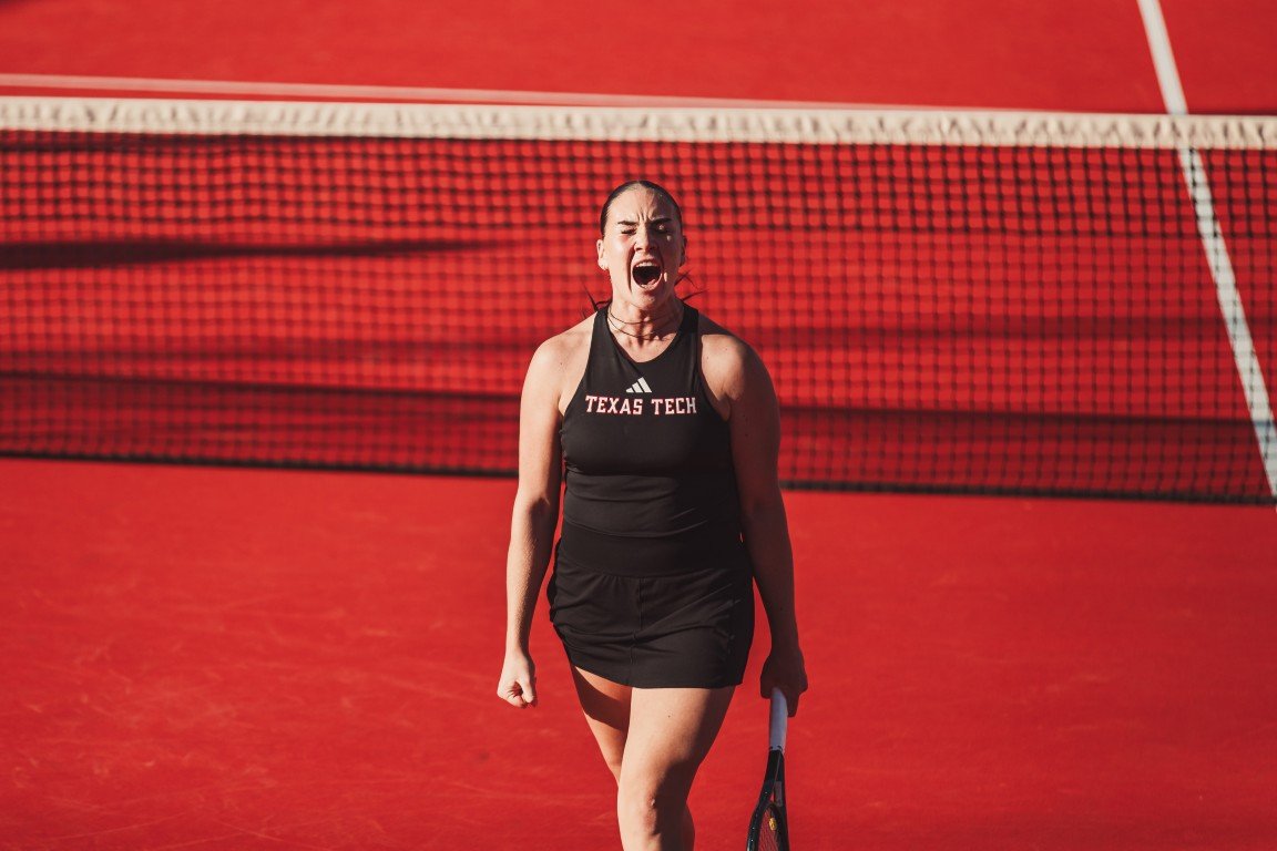 A female tennis player wearing a black sports outfit with 'TEXAS TECH' on the front, standing on a red tennis court with a net behind her, and appears to be shouting or celebrating.