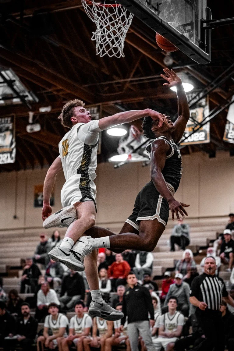 Two basketball players contest a shot at the basket during a game, with spectators in the background.