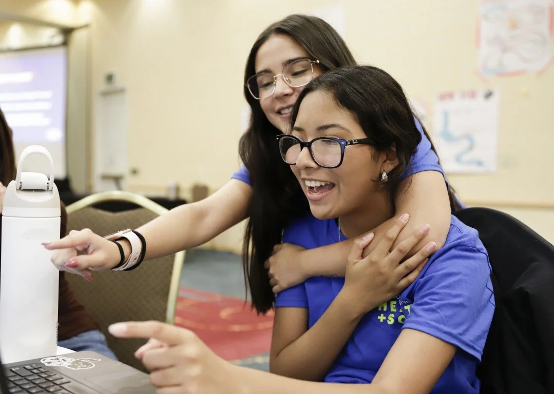 Two young women, one with dark hair and glasses, and the other with long dark hair, are smiling and sitting close together, looking at a laptop screen, in a room with a beige wall and a patterned carpet.