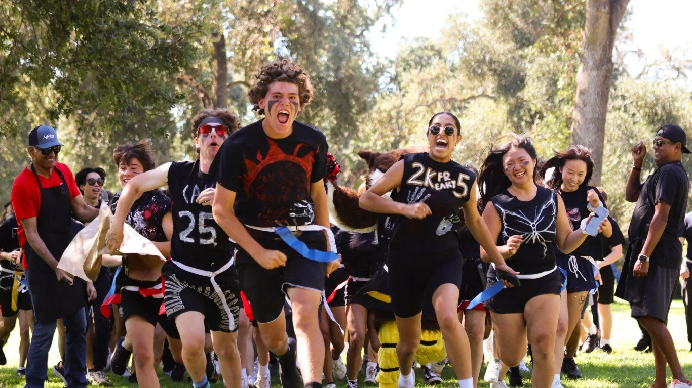 Group of people running outdoors in a park, celebrating and smiling, with some wearing costumes and sport-themed outfits.