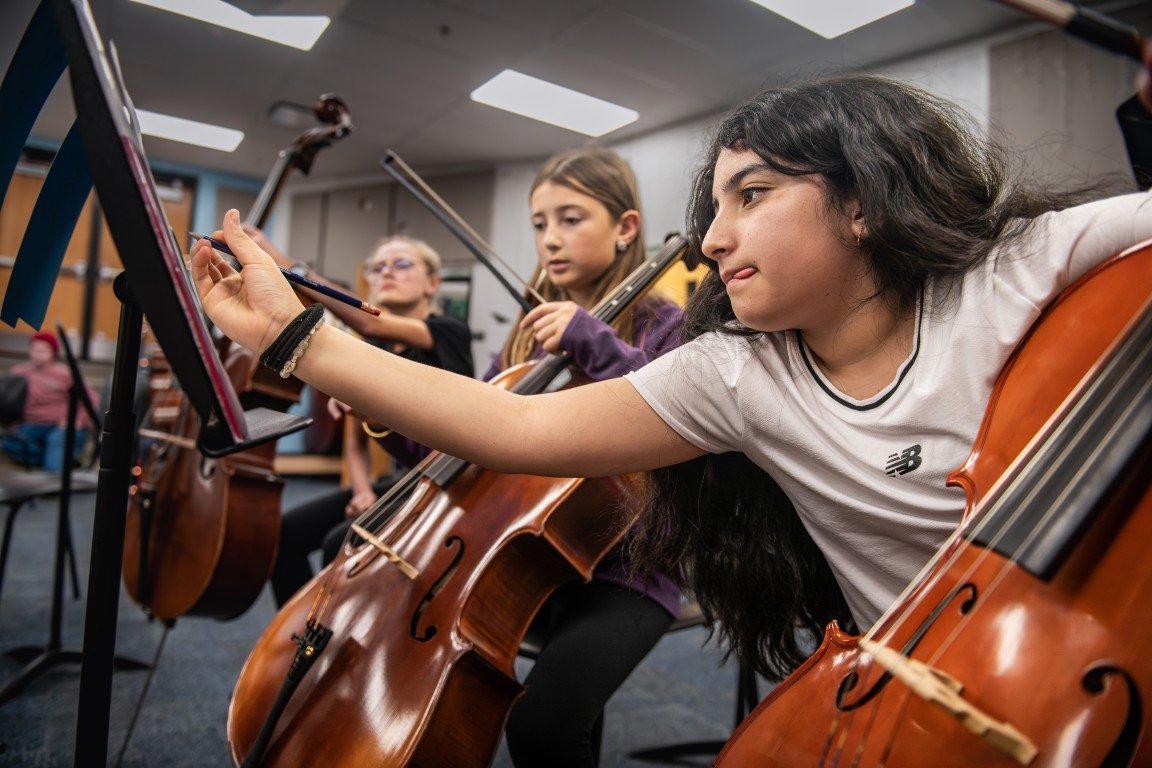 A girl in a white t-shirt adjusting her music stand while playing a cello in an orchestra rehearsal with other young musicians.
