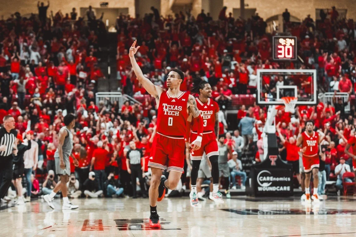 A Texas Tech basketball player in a red uniform with number 0 raising his hand after a game, surrounded by teammates and fans in a crowded arena.